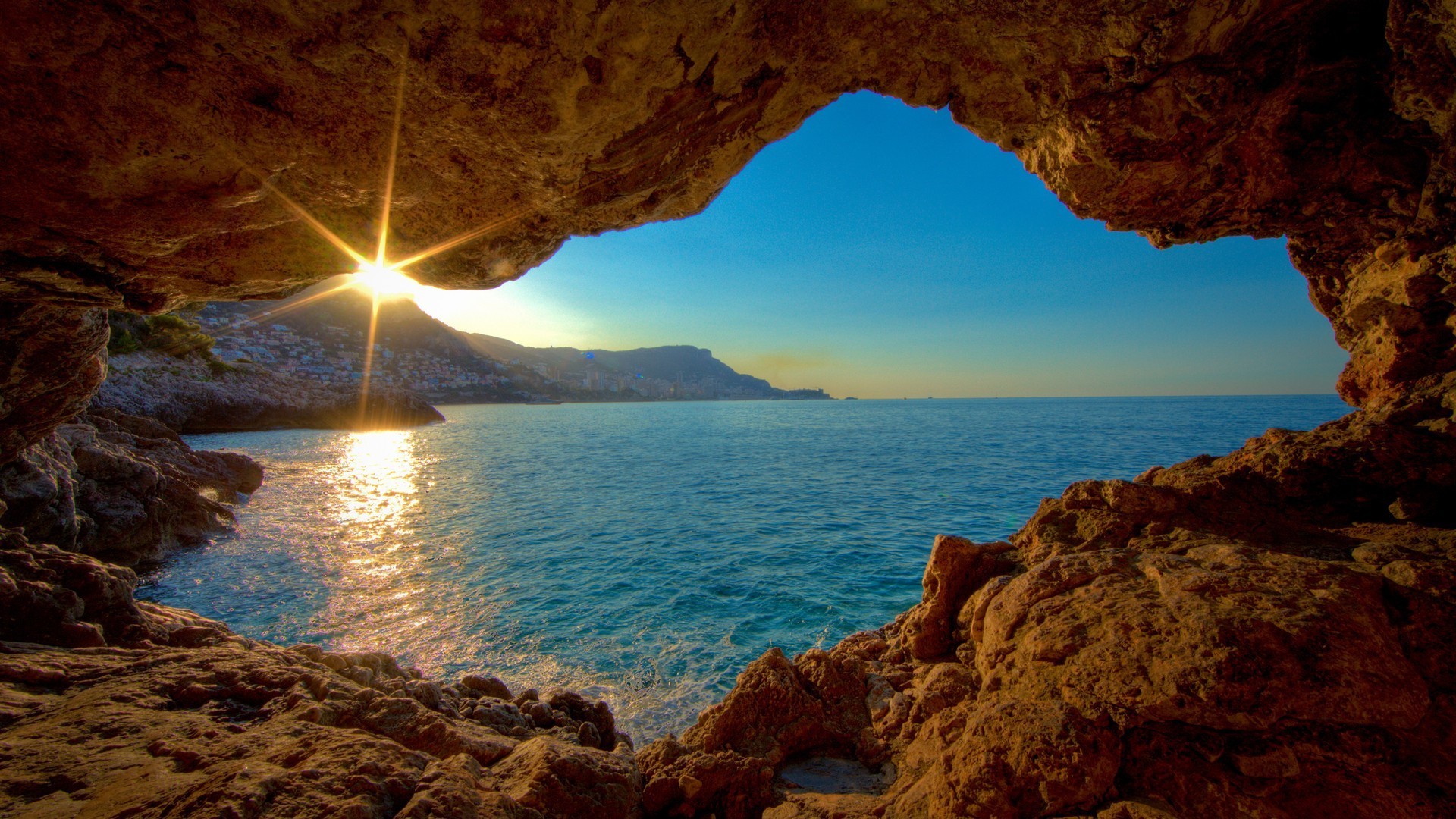 A breathtaking view of the ocean from inside a sea cave at sunset. The warm glow of the setting sun illuminates the rocky cave walls and the calm blue water, creating a serene and captivating scene