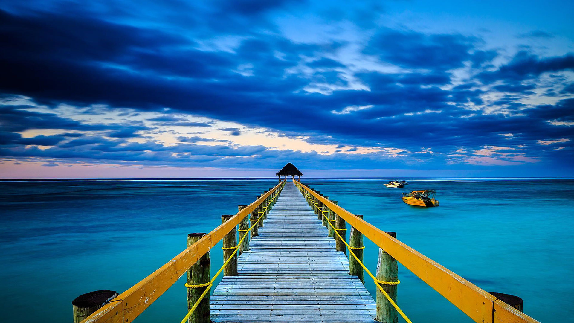 A long wooden pier stretches out into a tranquil turquoise ocean under a dramatic, cloud-filled sky. Two small yellow boats are anchored near the pier, adding a touch of color to the serene scene
