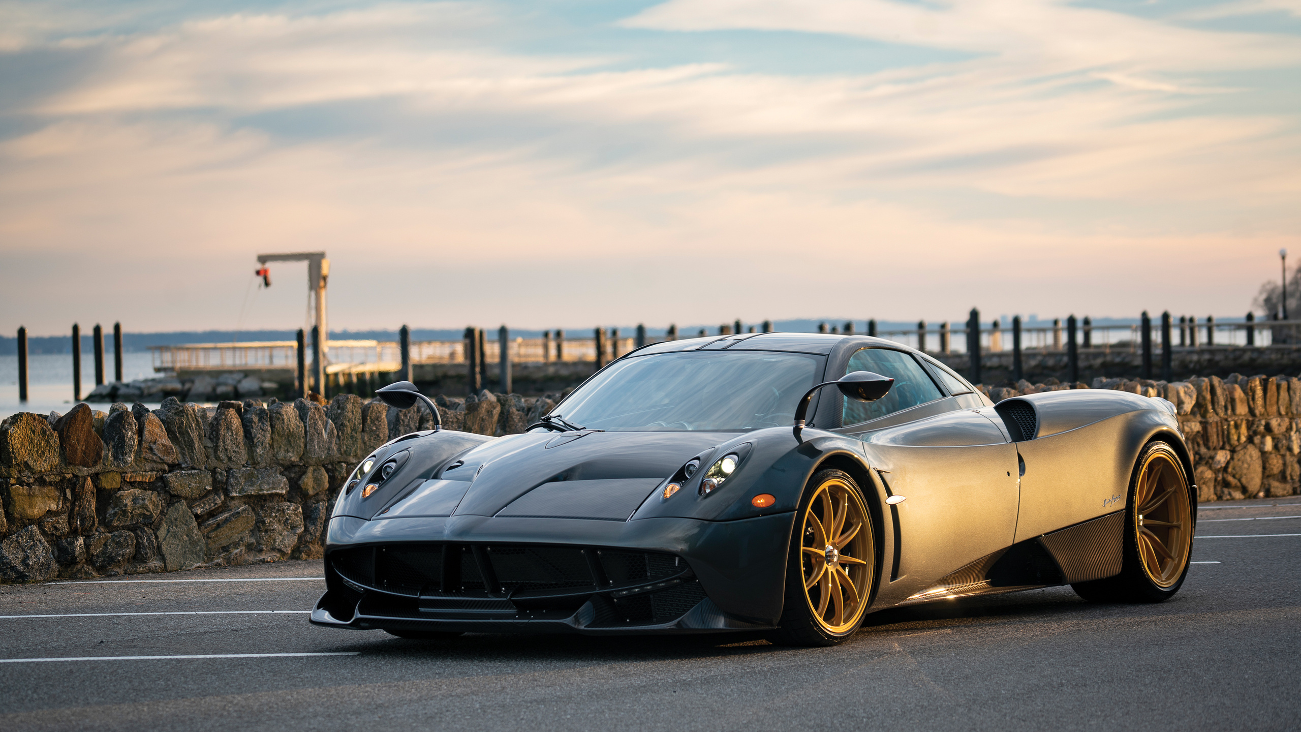 A sleek, dark gray Pagani Huayra supercar parked by the waterfront, showcasing its elegant design and gold-colored wheels
