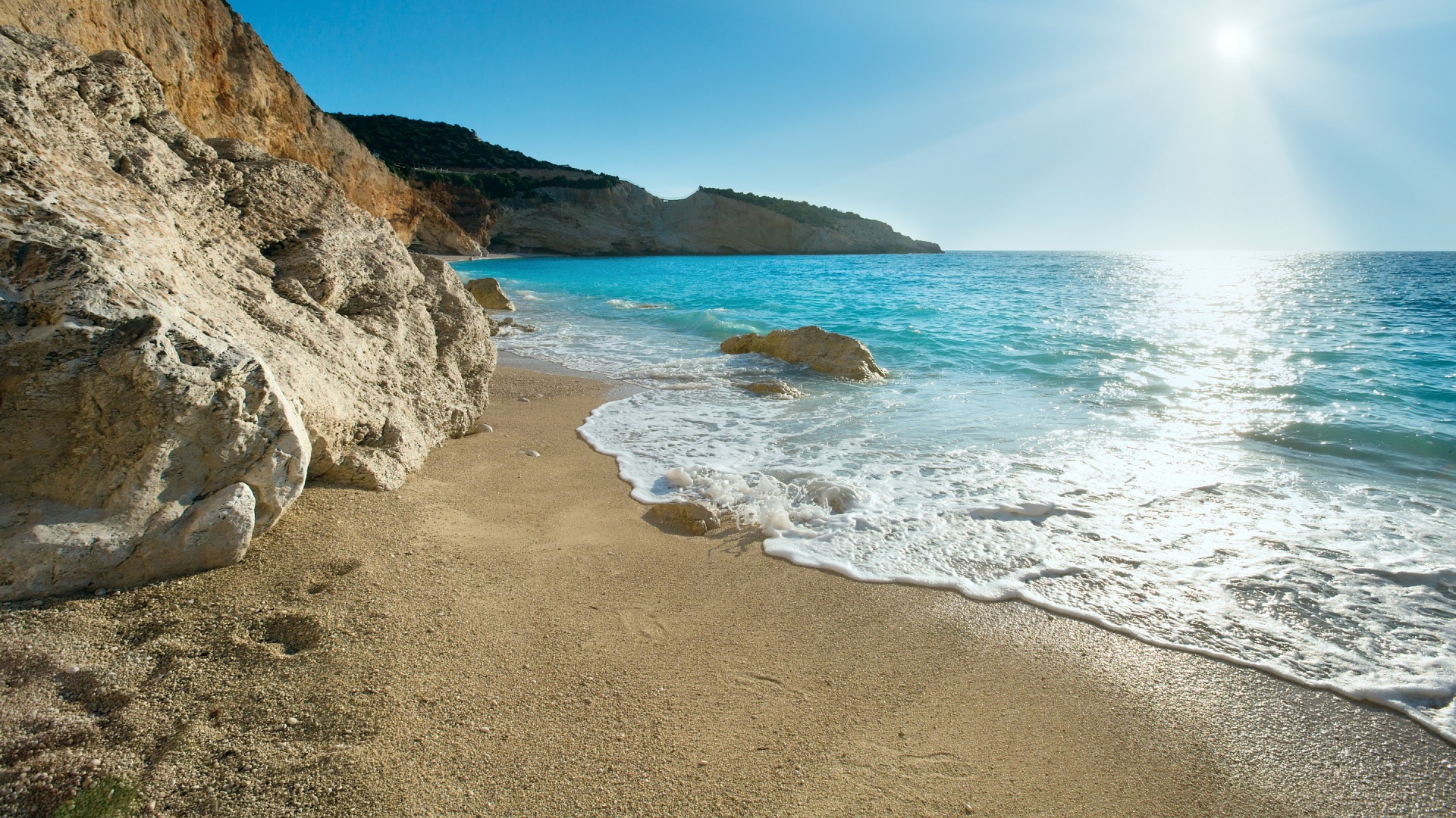 White sand beach on the Greek island meeting turquoise waters under a clear blue sky with the sun in the top right