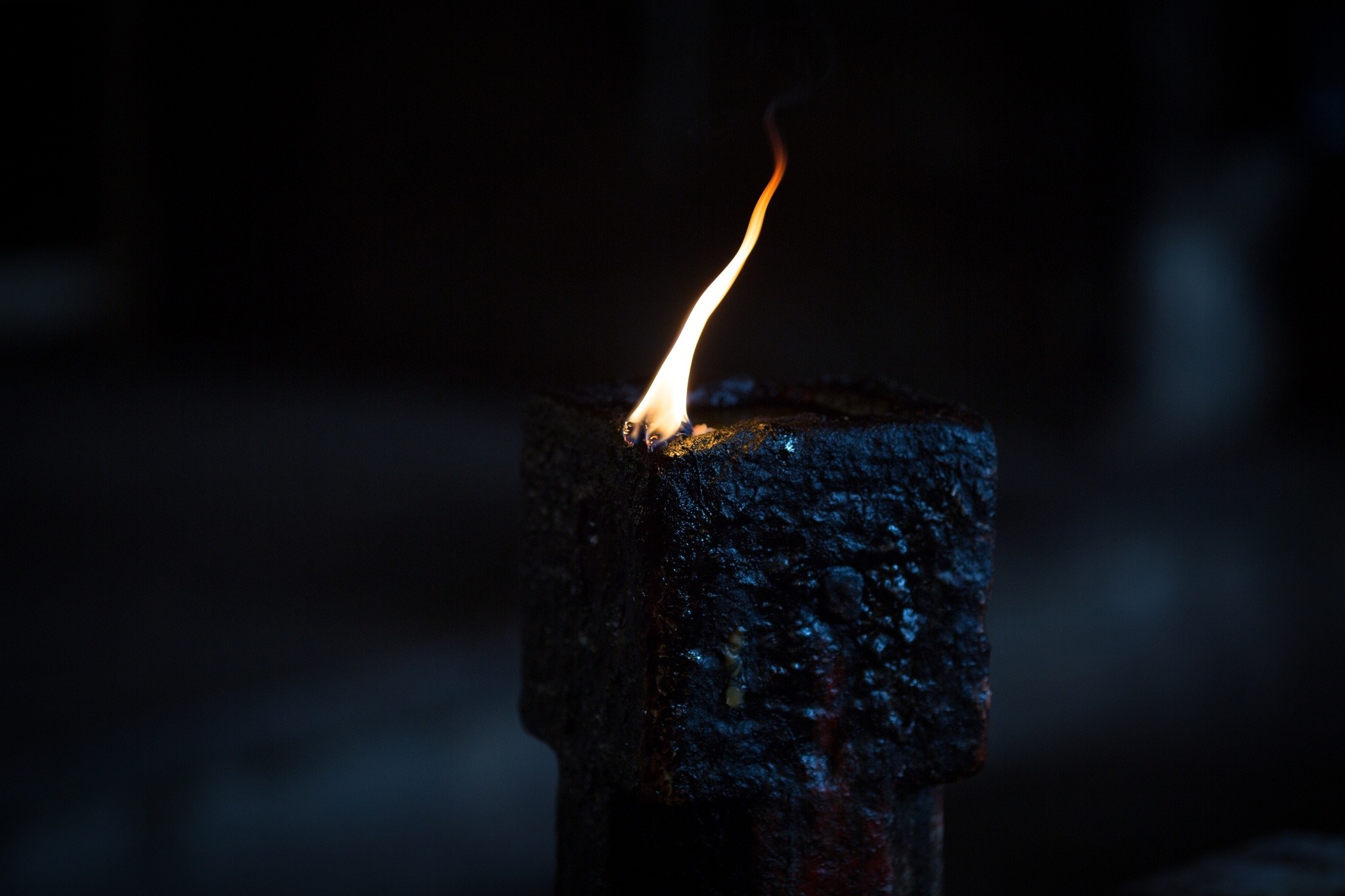 A single, flickering flame in a dark, textured oil lamp. The warm glow of the fire contrasts with the rough, dark surface of the lamp, creating a sense of peace and reverence