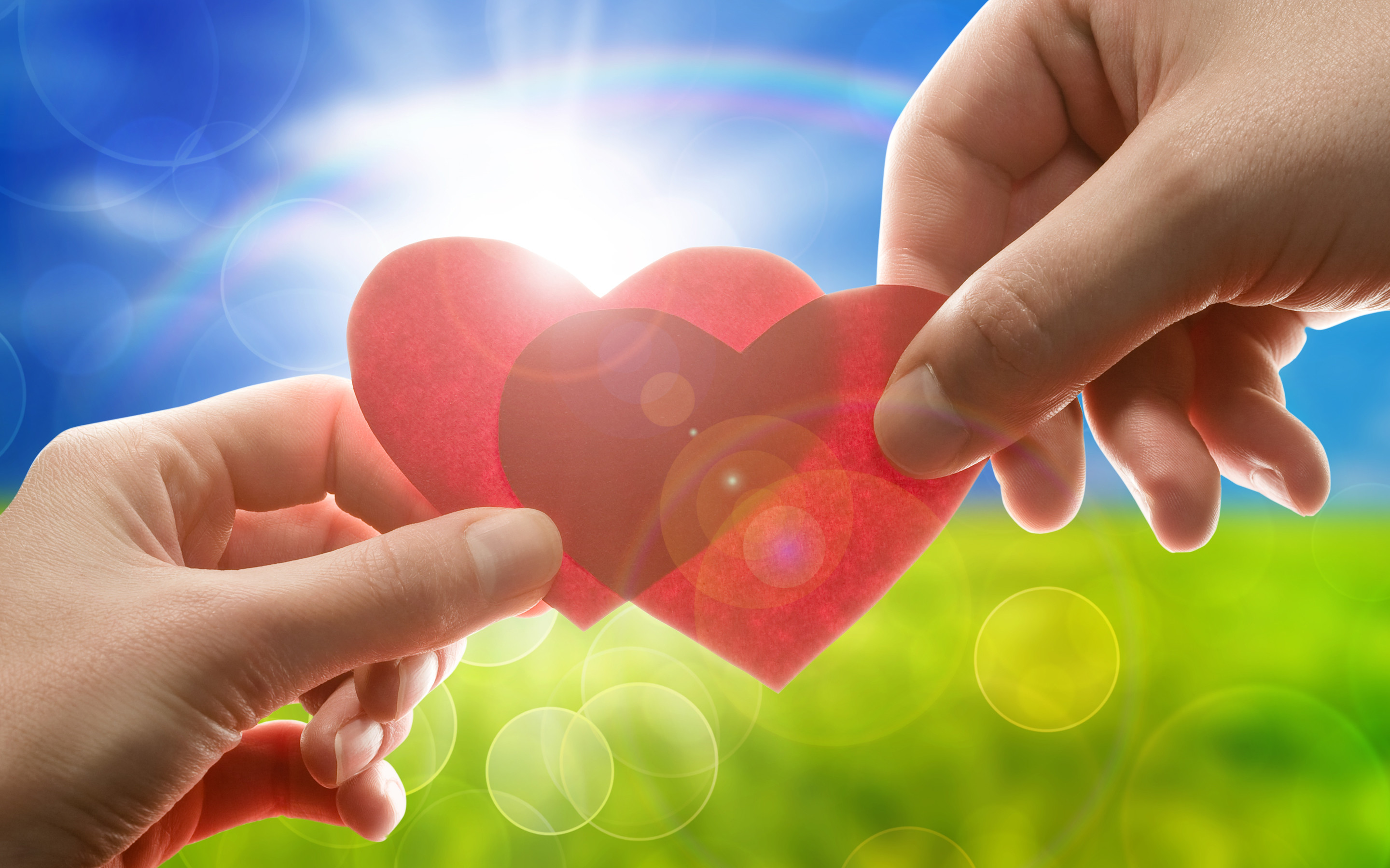 Two hands gently holding two red hearts against a backdrop of a sunny, green field, symbolizing friendship, love, and care