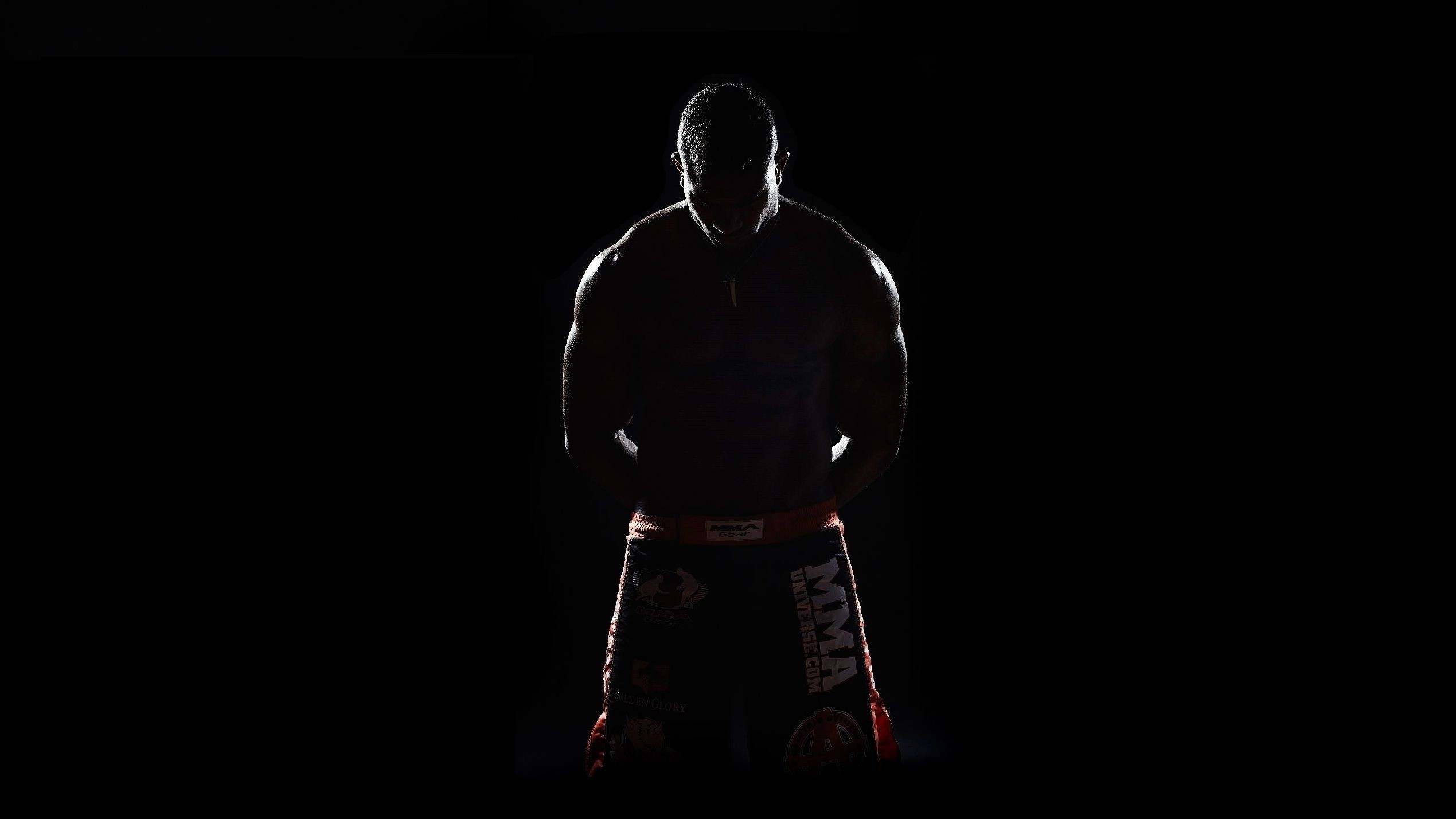 A silhouette of a muscular martial artist, likely an MMA fighter, stands with hands clasped in front, head bowed, against a dark background. The faint light outlines his physique and highlights the MMA Universe logo on his shorts