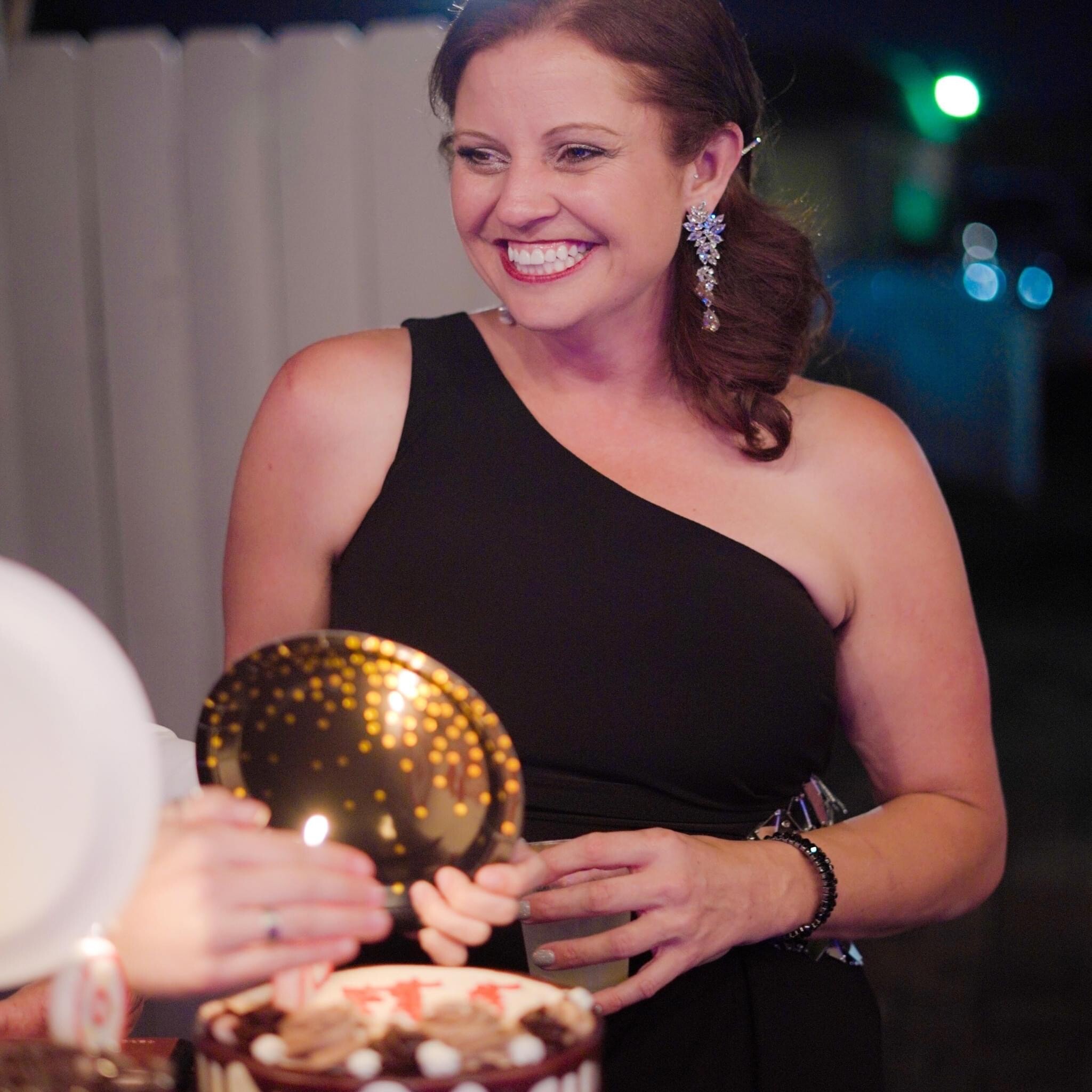 A woman in a black one-shoulder dress smiles at her birthday cake as someone lights a candle