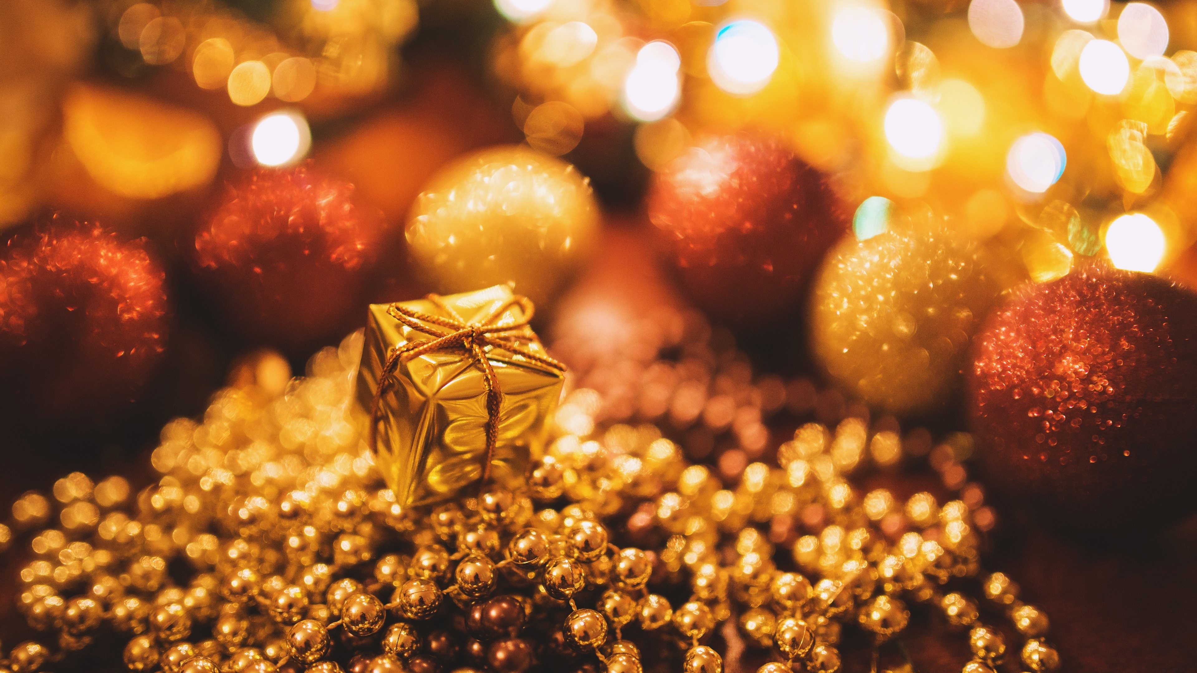 A close-up view of Christmas decorations, featuring a small, gold gift box resting on a bed of shimmering gold beads.  Red and gold ornaments, blurred by bokeh from festive lights, create a warm and inviting Christmas backdrop