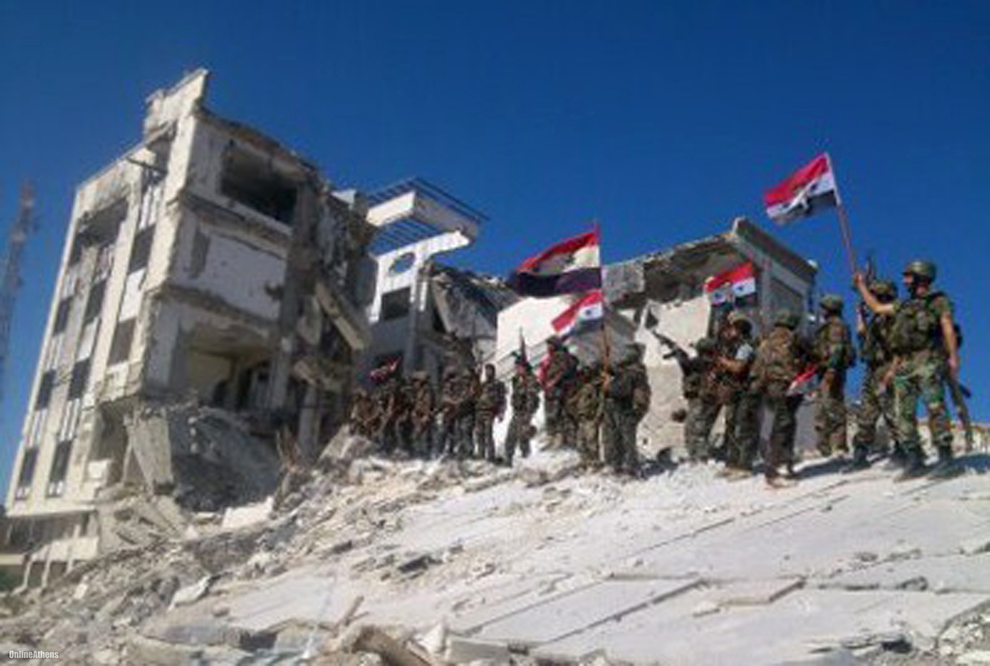 Syrian soldiers stand amidst the rubble of a destroyed building, holding Syrian flags, symbolizing the impact of the Syrian Civil War