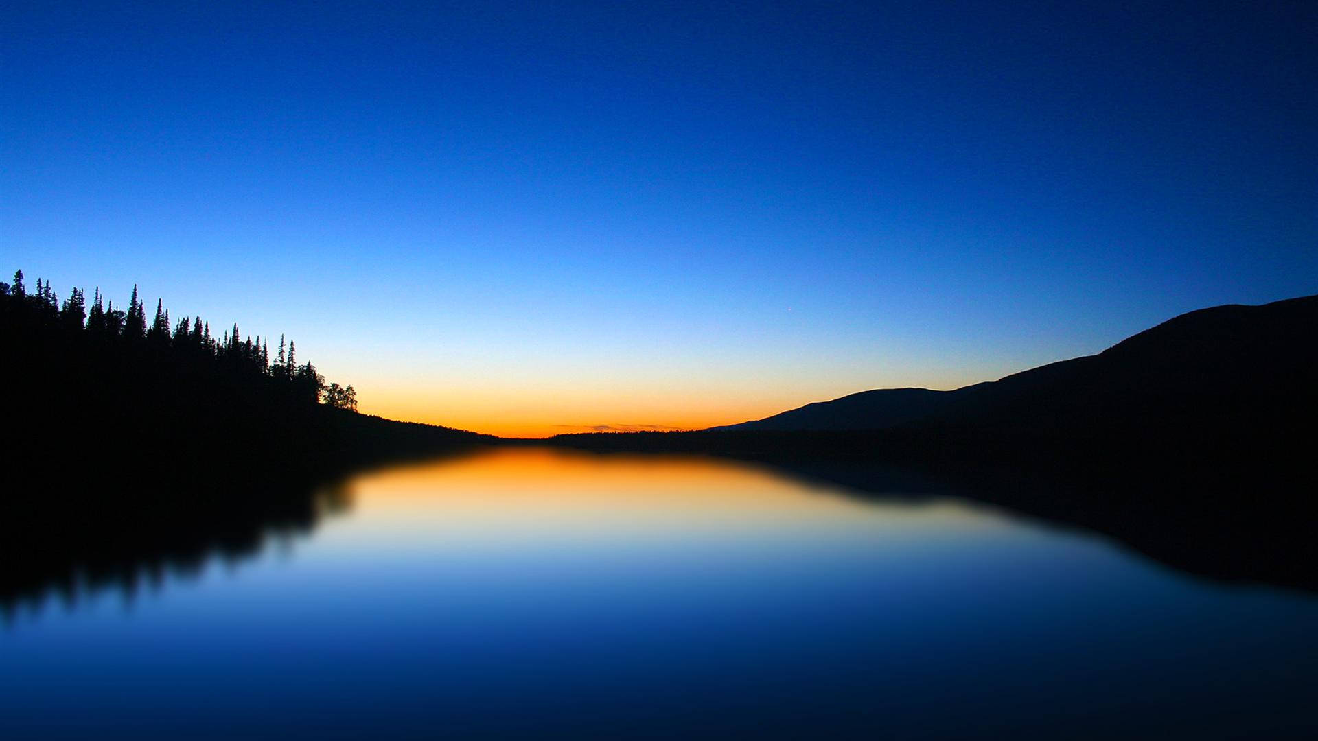 A serene sunset over a calm lake, with silhouetted trees and mountains in the background