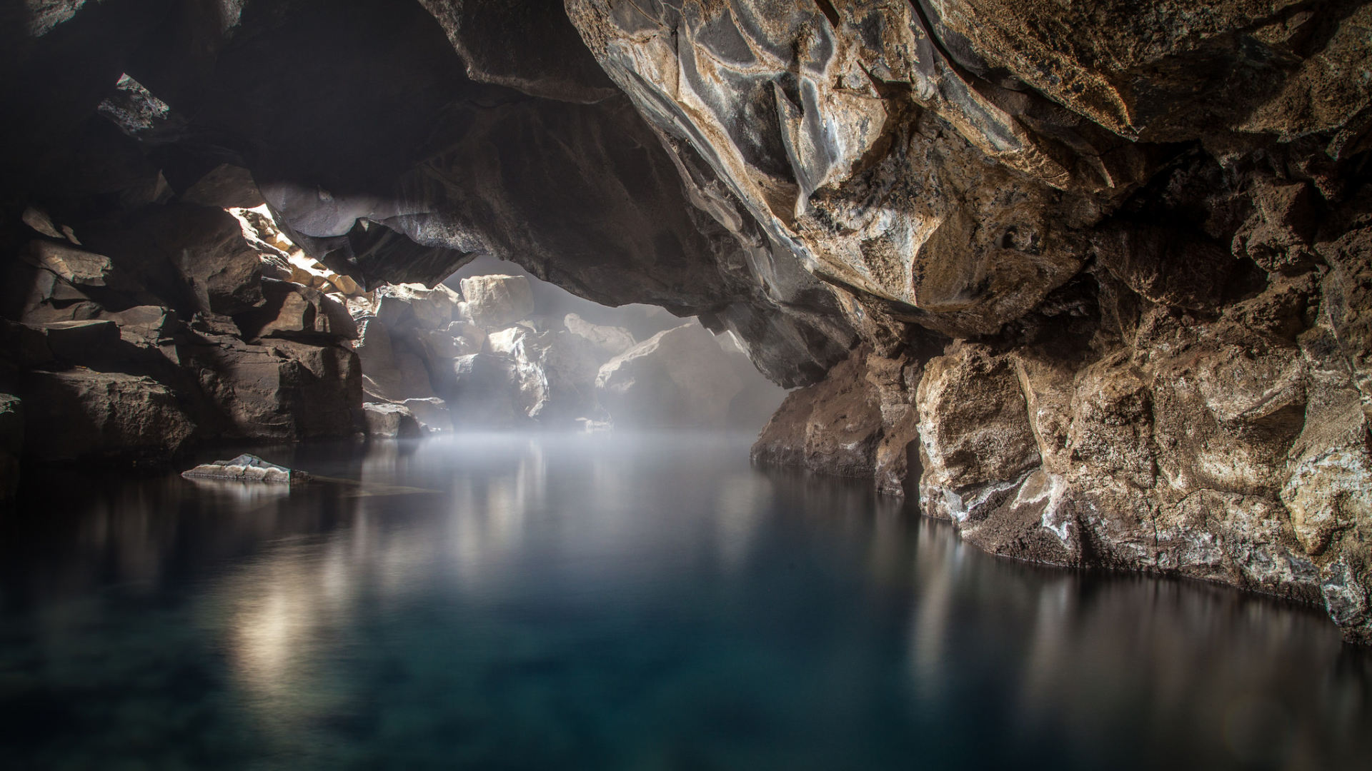 A mesmerizing view of a cave with a subterranean pool reflecting the light filtering through the cave opening. The ethereal mist adds to the mystical ambiance of this natural wonder