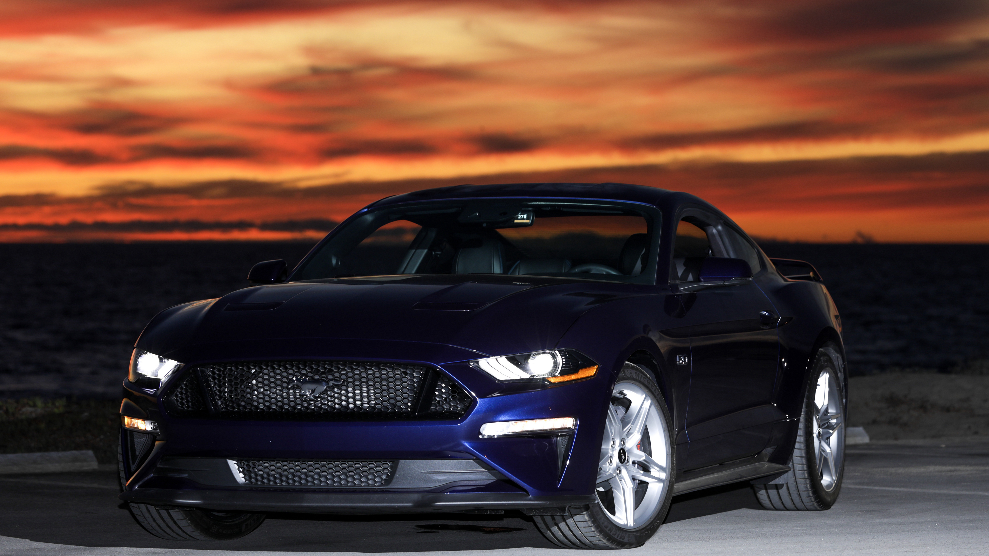 A dark blue Ford Mustang parked by the ocean at sunset. The vibrant colors of the sky reflect off the car's sleek body, creating a stunning image