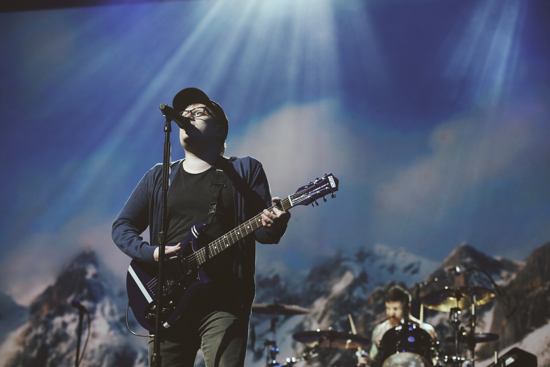 Patrick Stump of Fall Out Boy performing live on stage, singing into a microphone and playing a dark-colored electric guitar. A drummer is visible in the background