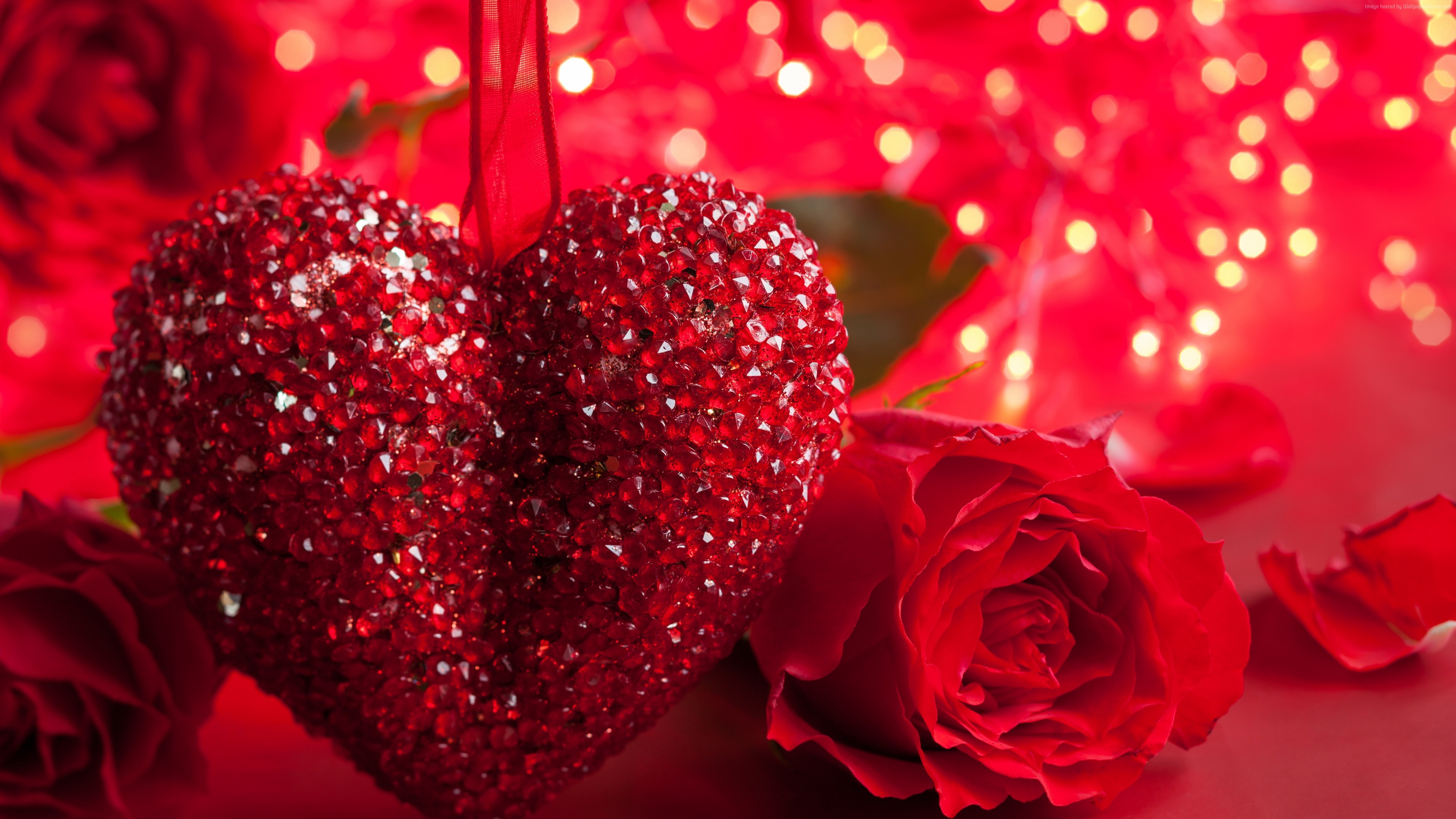 A sparkling red heart ornament hangs amidst red roses, illuminated by a warm, romantic glow of string lights, creating a perfect Valentine's Day scene