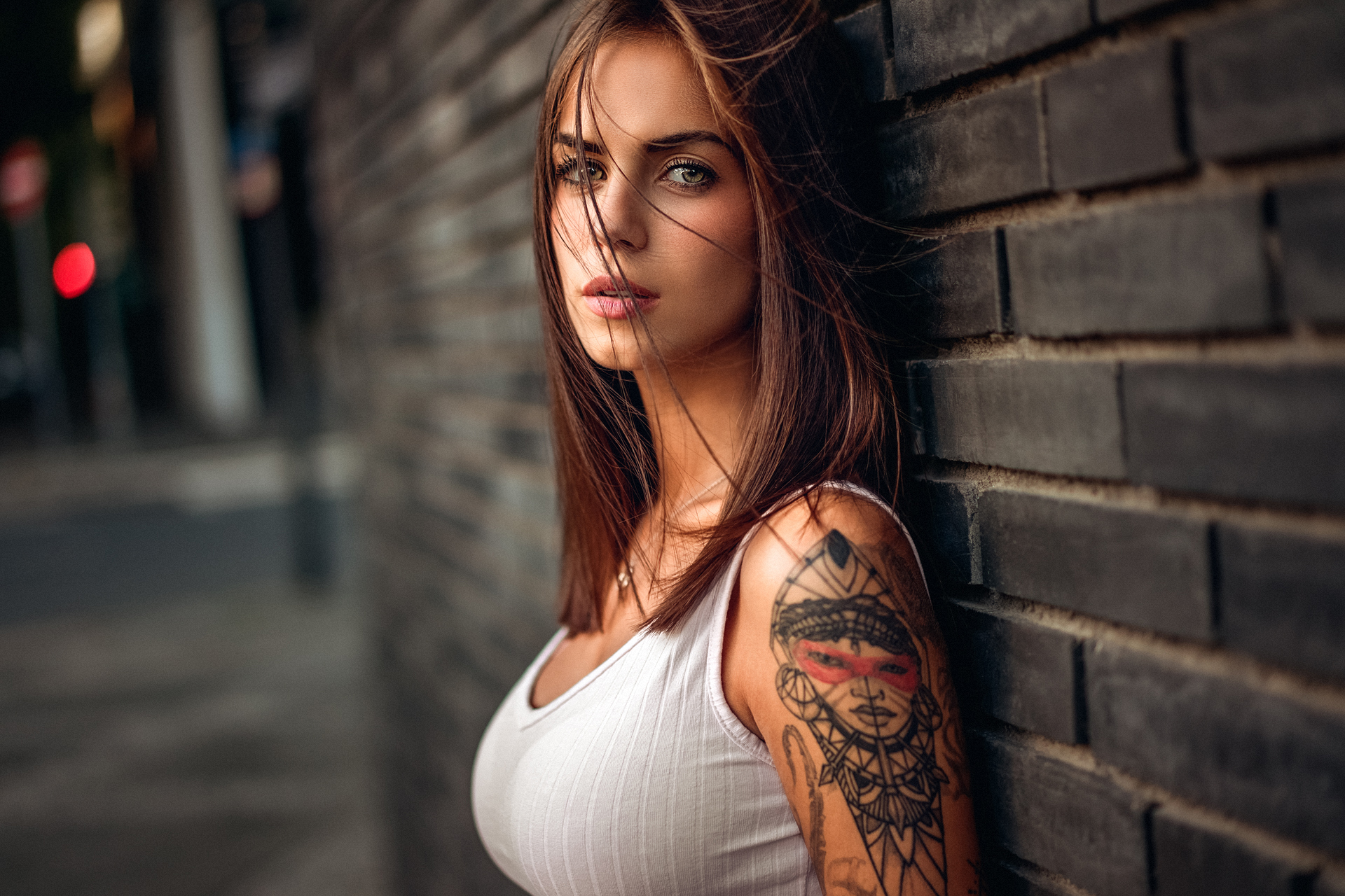 A young woman with long brown hair and a tattoo on her arm leans against a brick wall