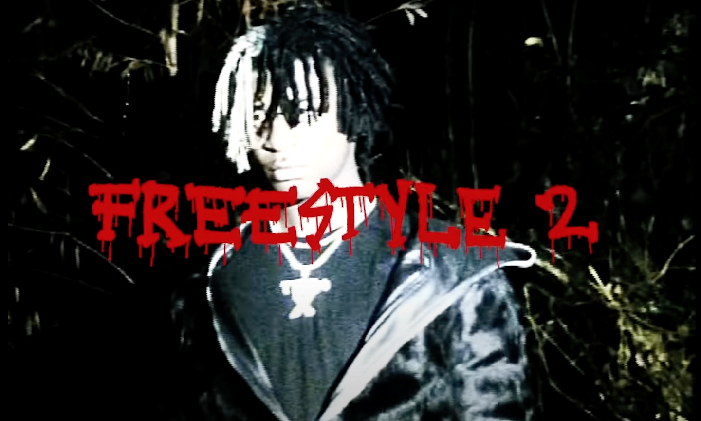 A person with two-toned dreadlocks, wearing a dark jacket and a cross necklace, stands against a blurred natural background. Red text overlaid on the image reads 'FREESTYLE 2'