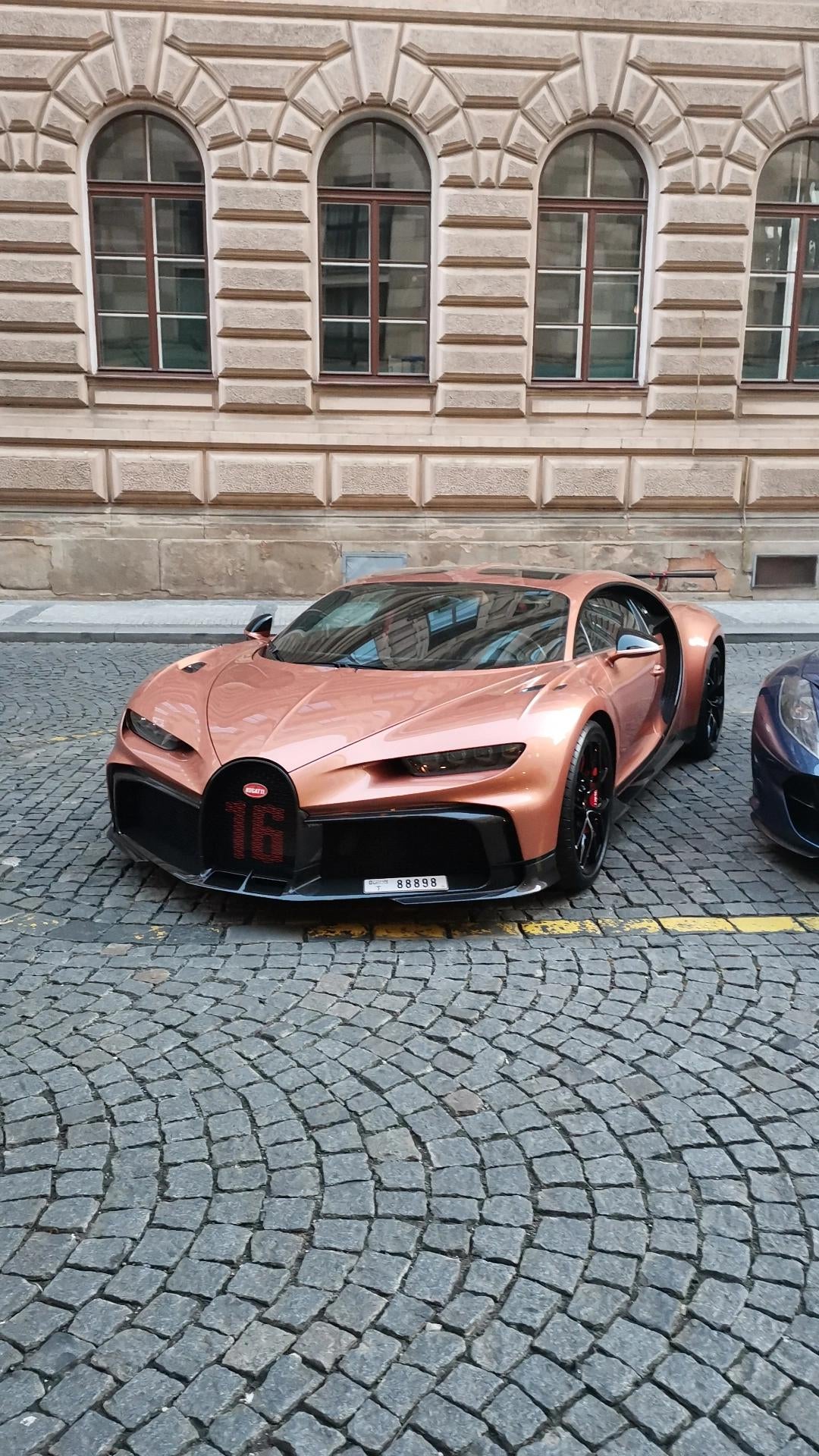 A rose gold Bugatti Chiron Pur Sport parked on a cobblestone street in front of a tan building
