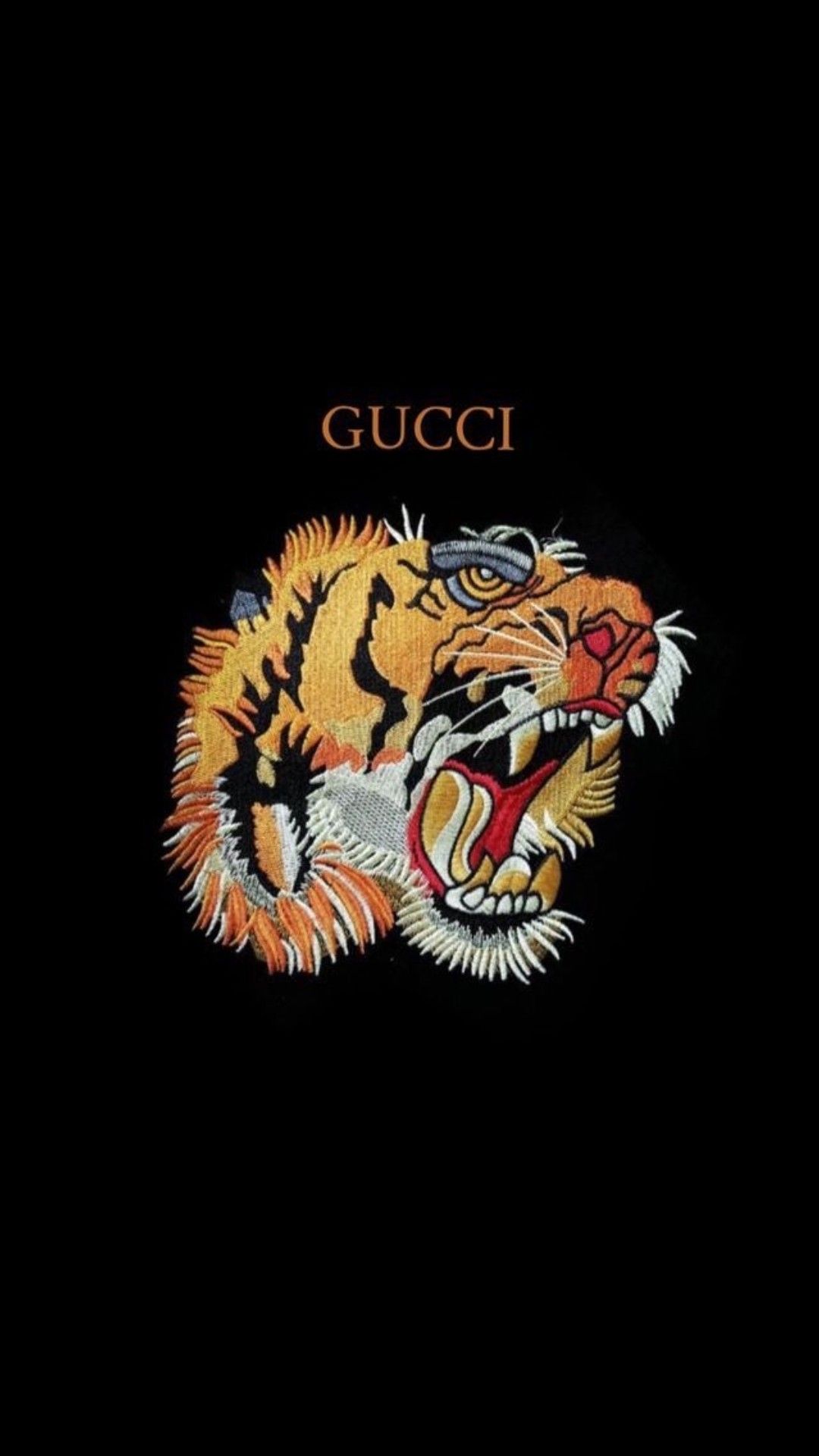 A close-up of a roaring tiger head, likely embroidered or printed on a black Gucci item, set against a stark black background. The tiger, a recurring motif in Gucci designs, is depicted with detailed orange and black stripes, white whiskers, and sharp teeth