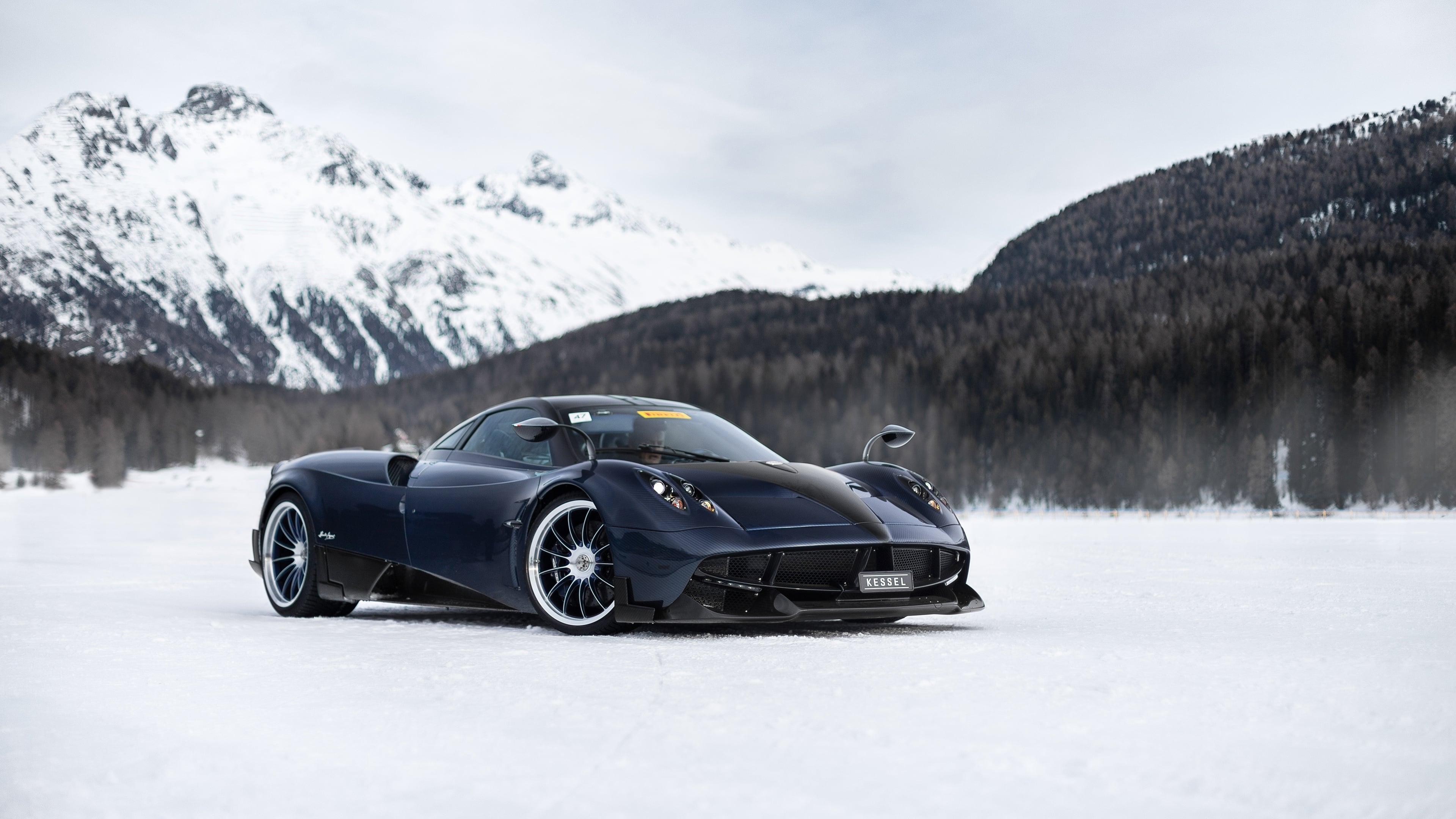 A dark blue Pagani Huayra sits on a snowy surface, with snow-covered mountains and a forest in the background