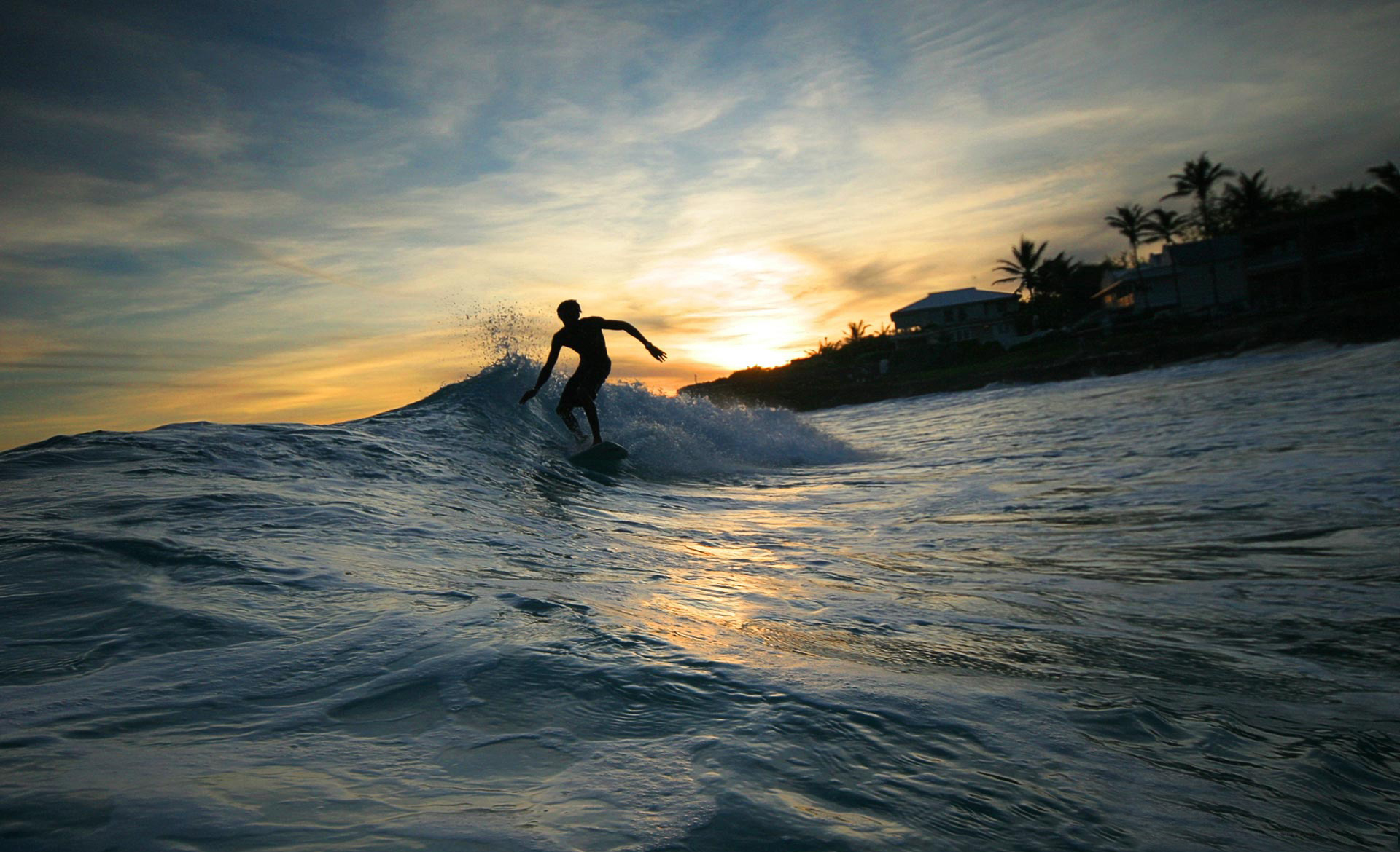 Silhouette of a surfer riding a wave at sunset with tropical coastline in background
