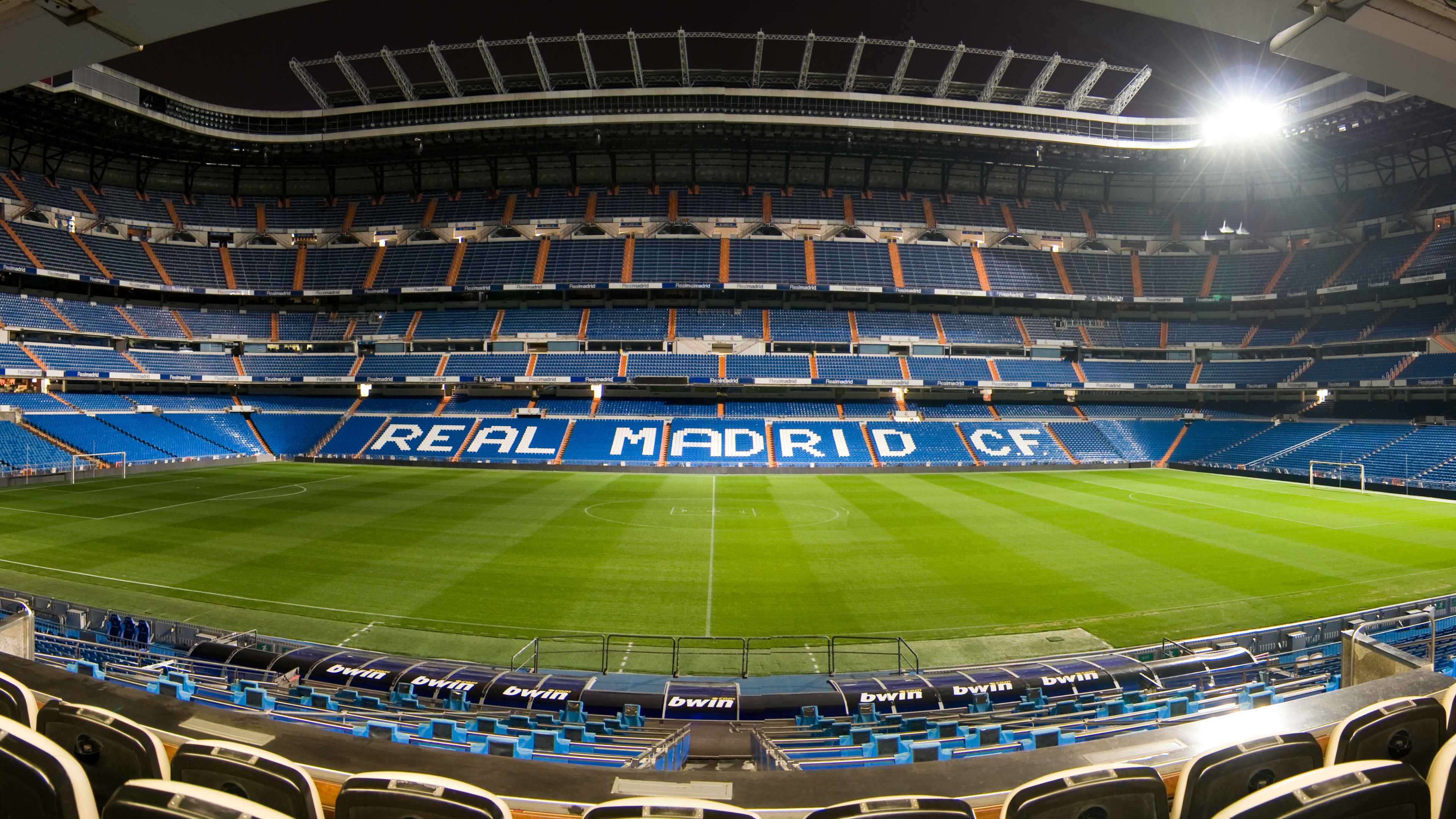 A panoramic view of the Santiago Bernabéu Stadium, home of Real Madrid, illuminated at night. The lush green field and blue seats create a striking contrast