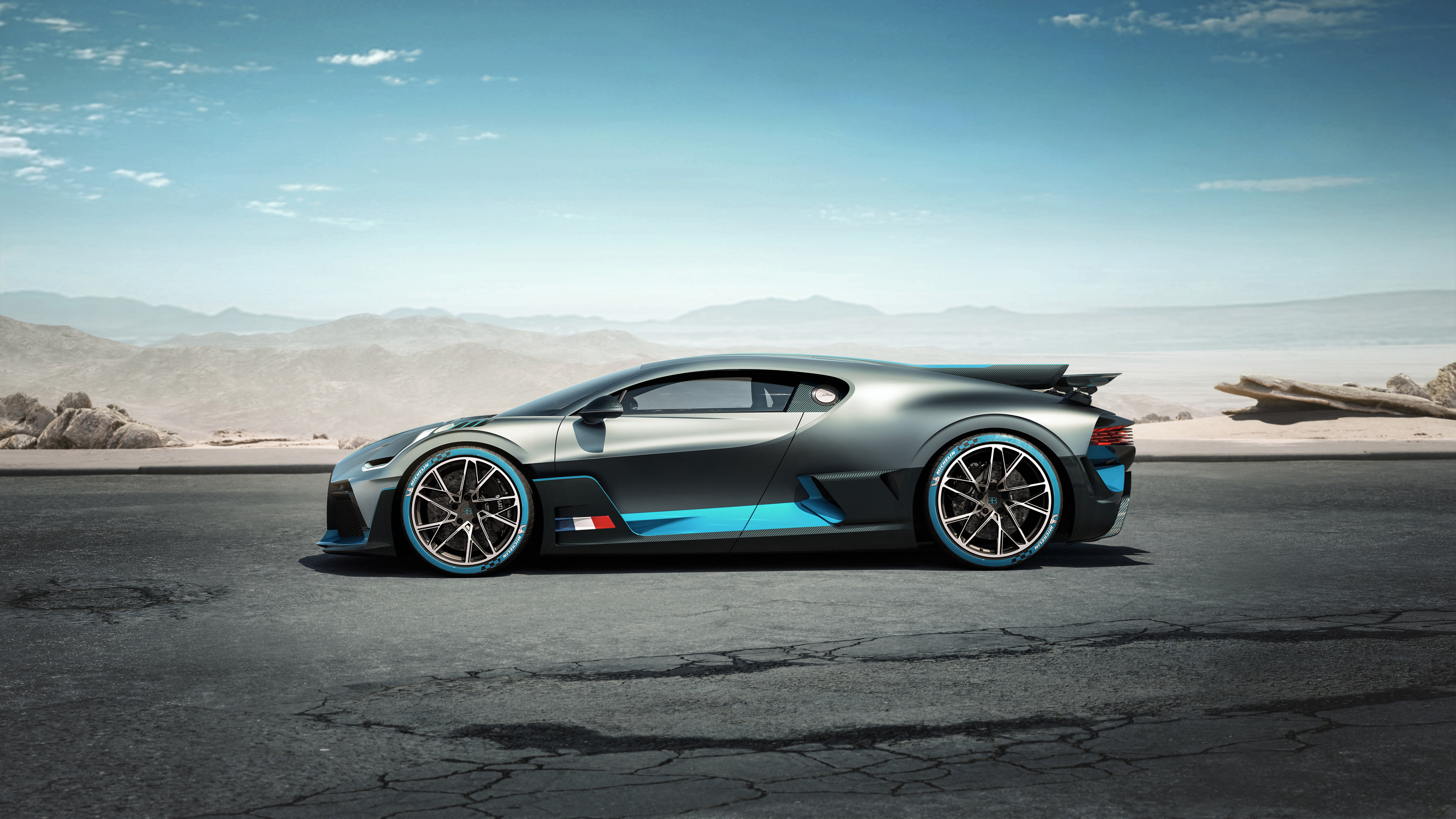 A sleek, futuristic Bugatti concept car, the Bolide, is showcased in profile against a desert backdrop. Its dark gray body is accented with vibrant blue racing stripes and matching blue highlights on the tires, emphasizing its speed and performance