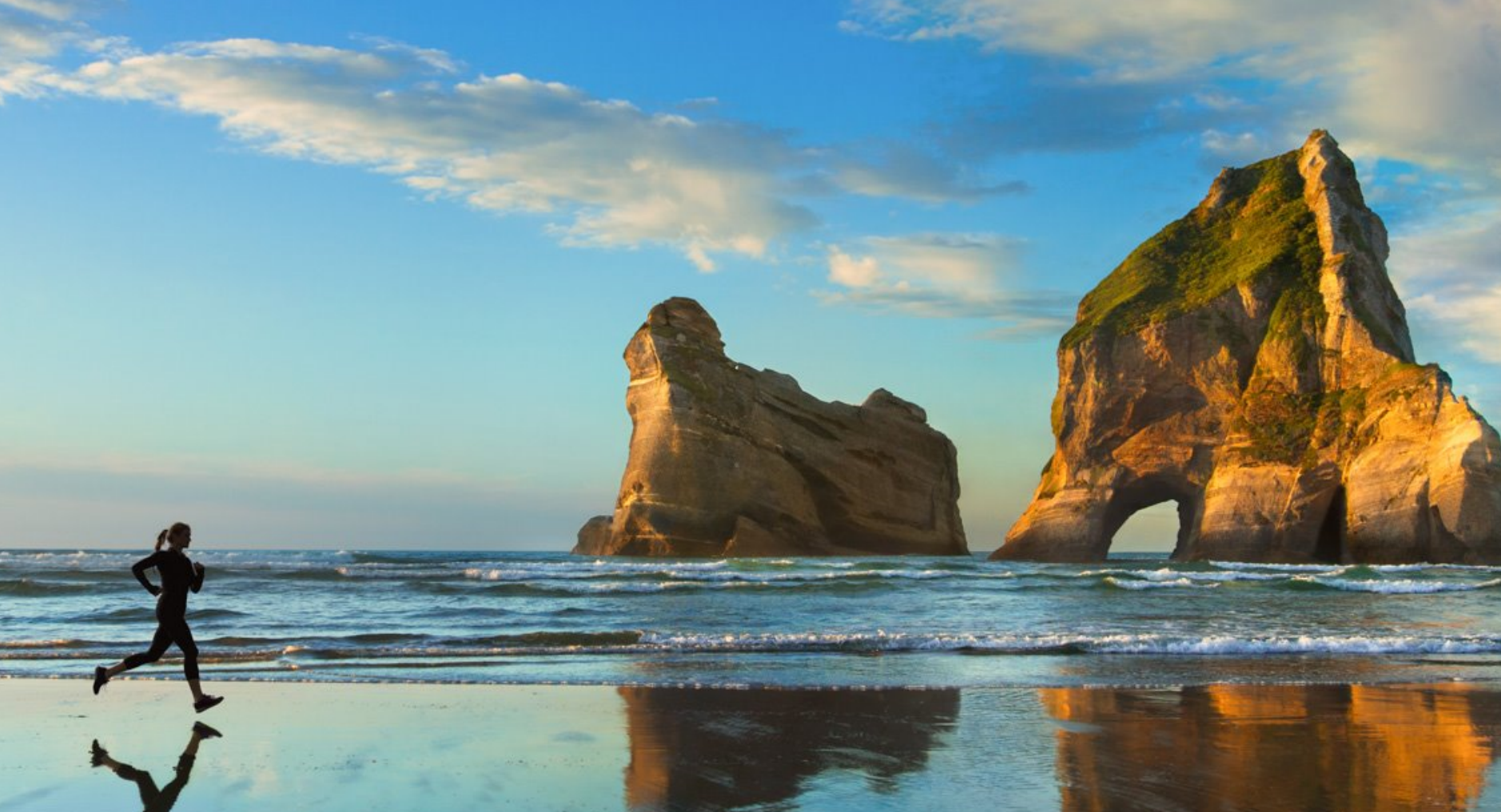 A lone runner on Wharariki Beach, New Zealand, with the iconic rock formations reflected in the wet sand at sunset