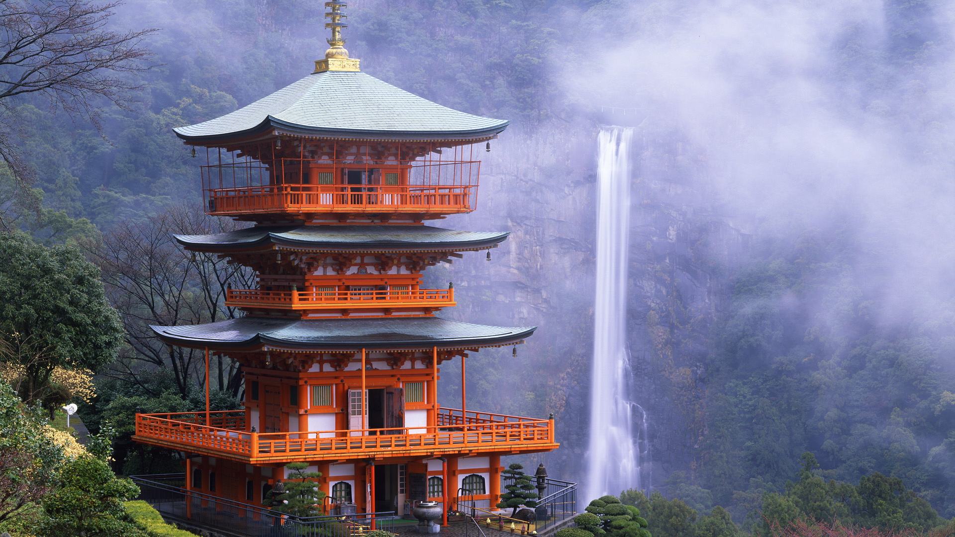 A vibrant orange pagoda stands majestically against the backdrop of a misty waterfall in Japan