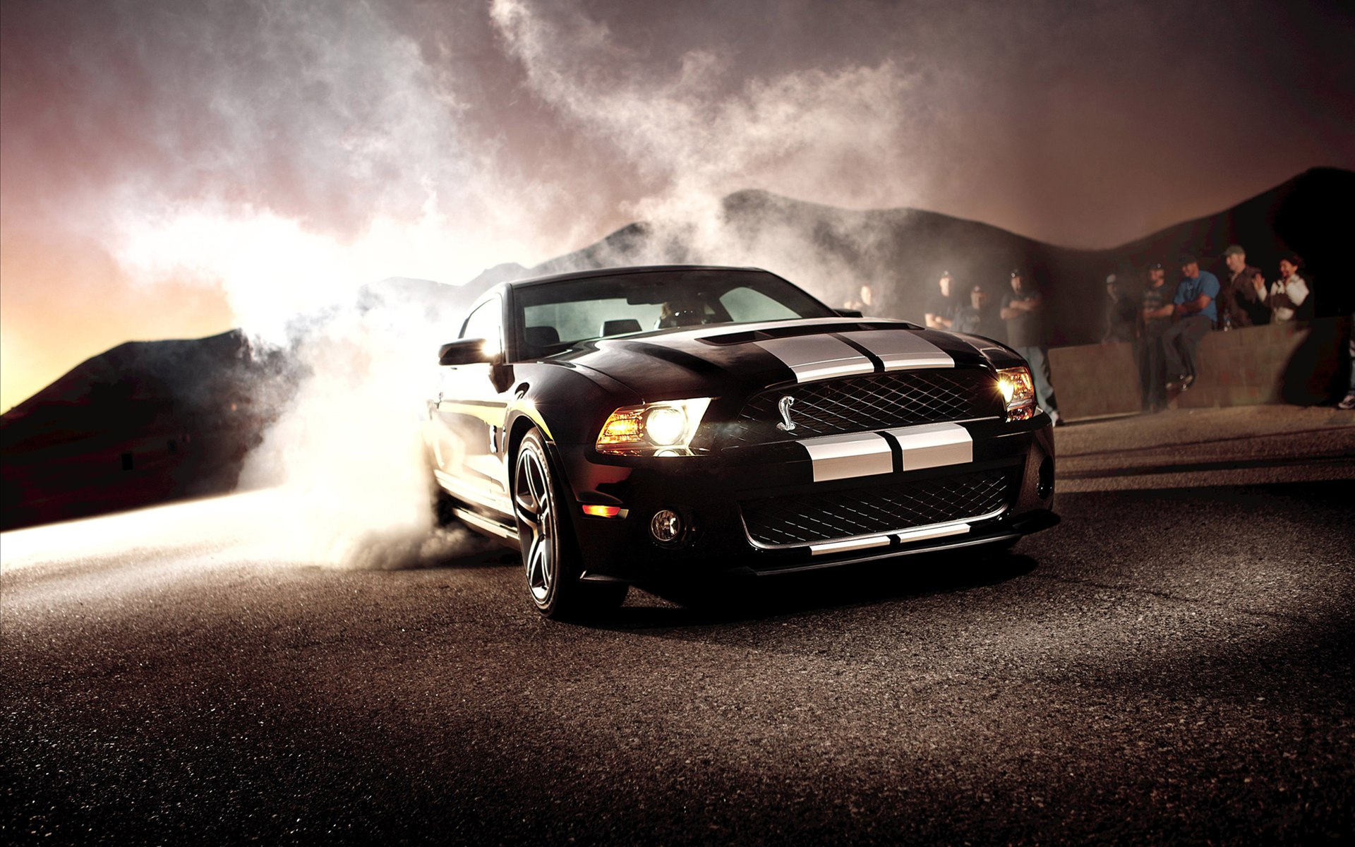 Black Ford Mustang Shelby GT500 with white racing stripes drifting on asphalt at dusk, surrounded by smoke, with spectators in the background
