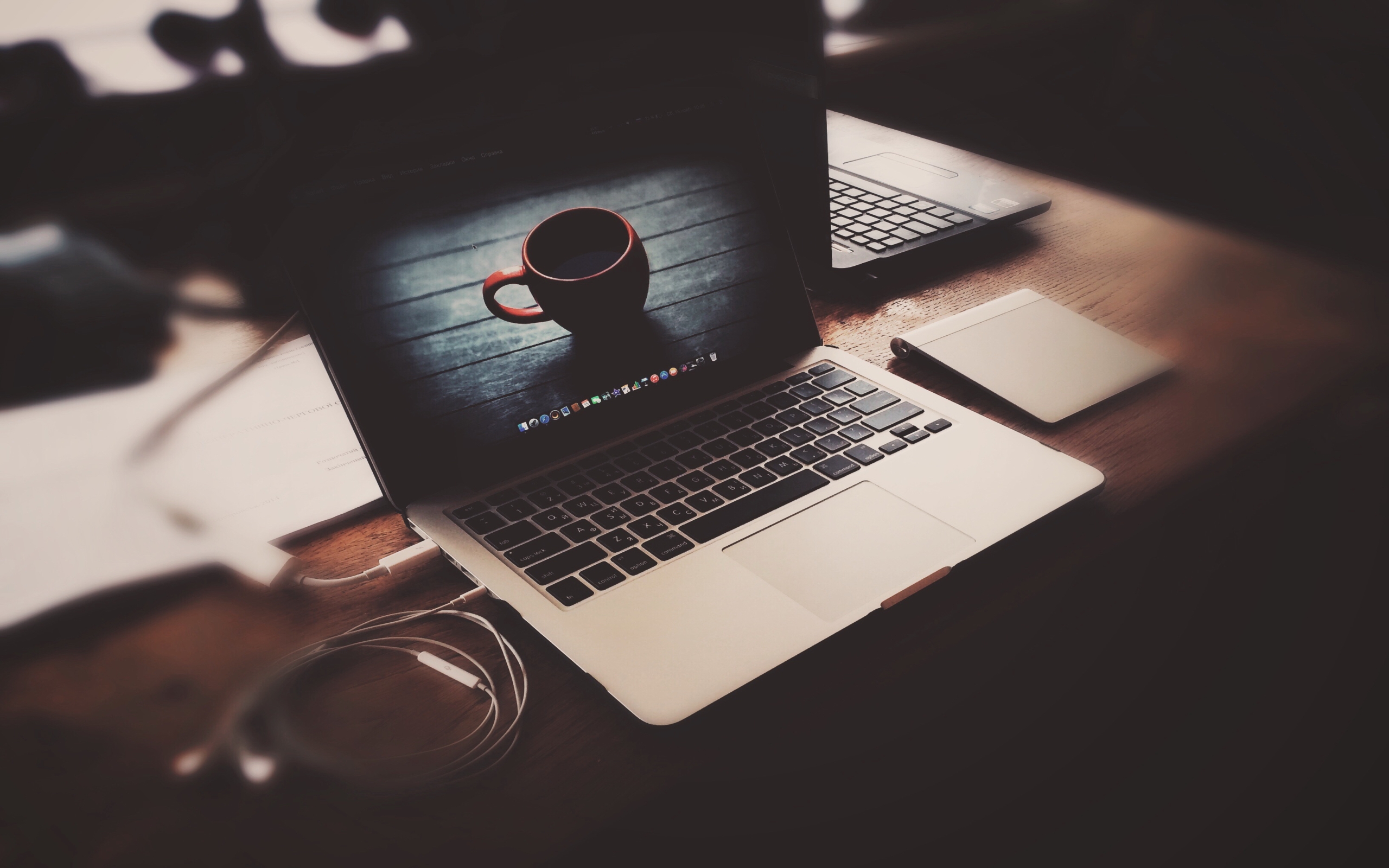 A silver MacBook laptop open on a wooden desk, displaying a wallpaper of a coffee cup on a wooden surface. A second laptop and a tablet are slightly blurred in the background
