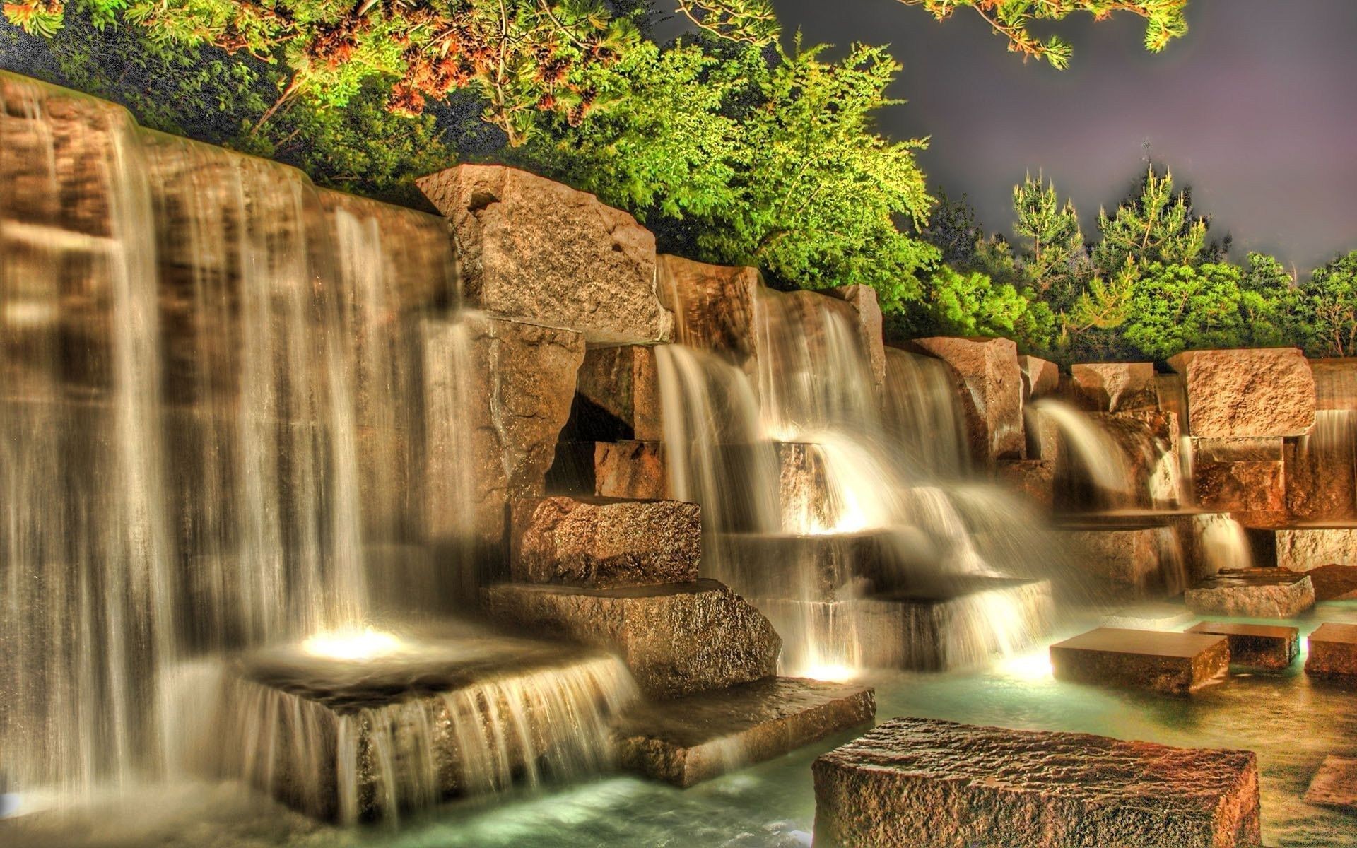 A mesmerizing long-exposure photograph of a multi-tiered waterfall cascading over illuminated rocks at night. The lush greenery in the background adds to the serene ambiance