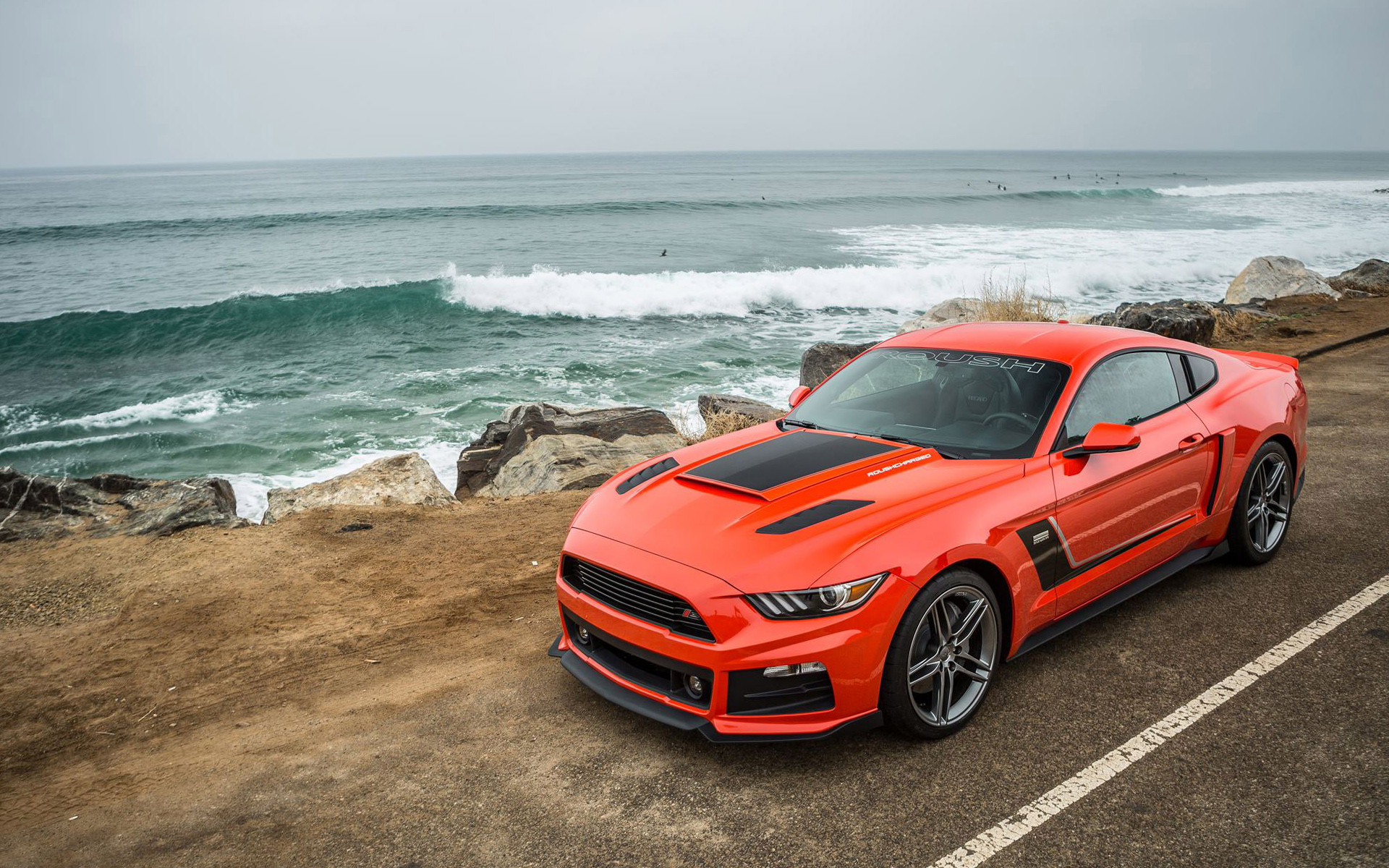 Orange Ford Mustang parked by the ocean with surfers in the background