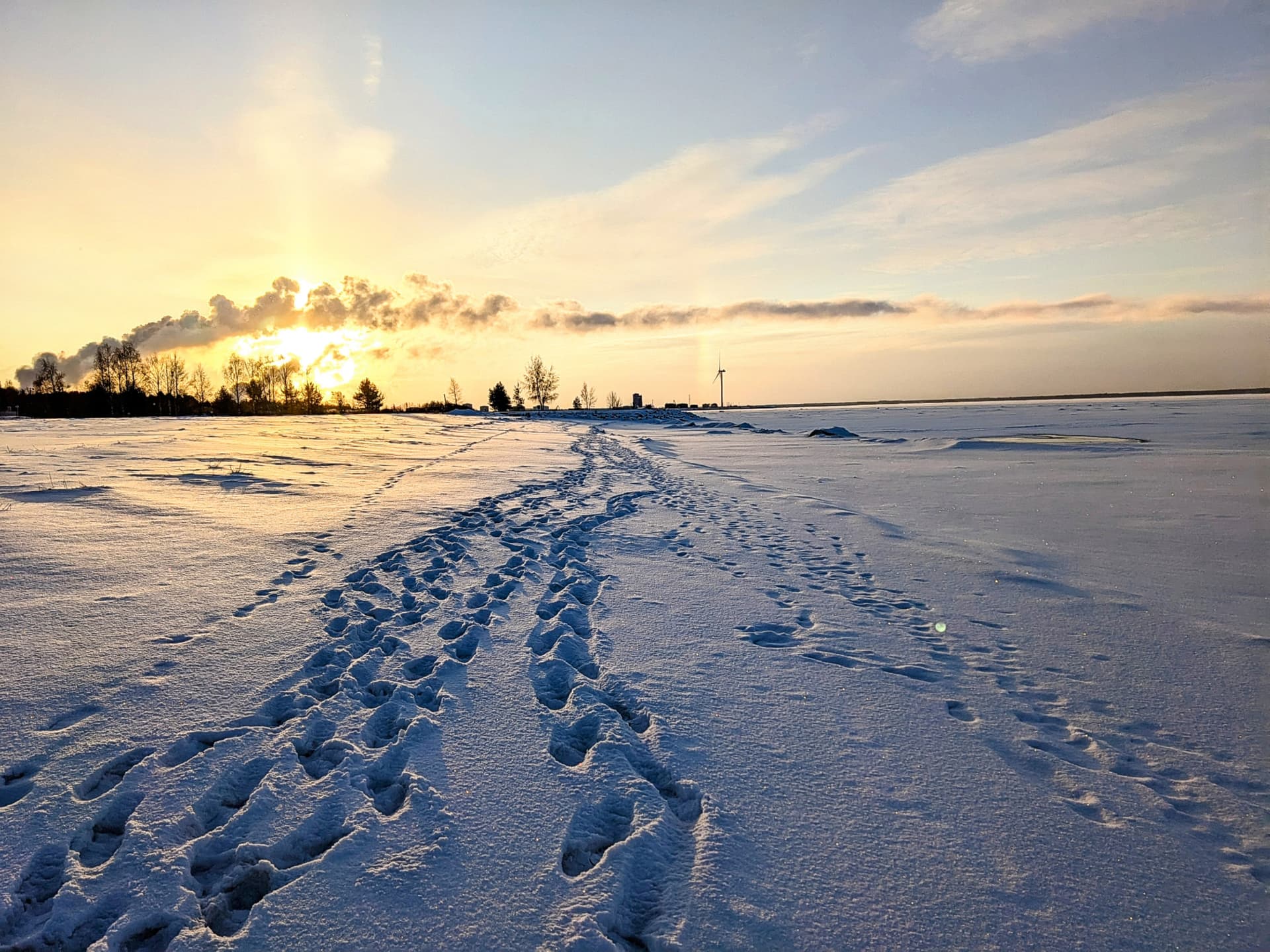 Footprints in the snow lead towards a frozen lake at sunset. A wind turbine and trees are visible in the distance