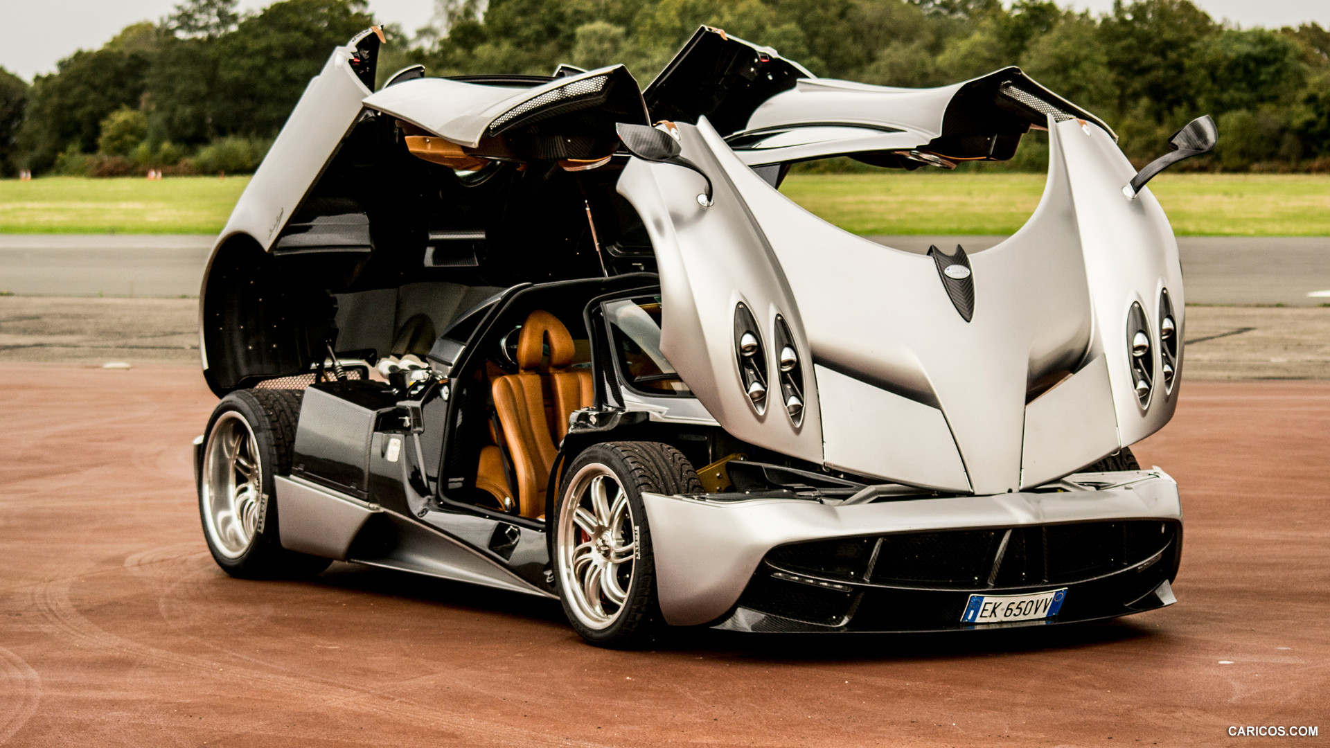 A silver Pagani Huayra with its gull-wing doors open, revealing the luxurious tan interior. The car is parked on a reddish-brown surface, possibly a race track