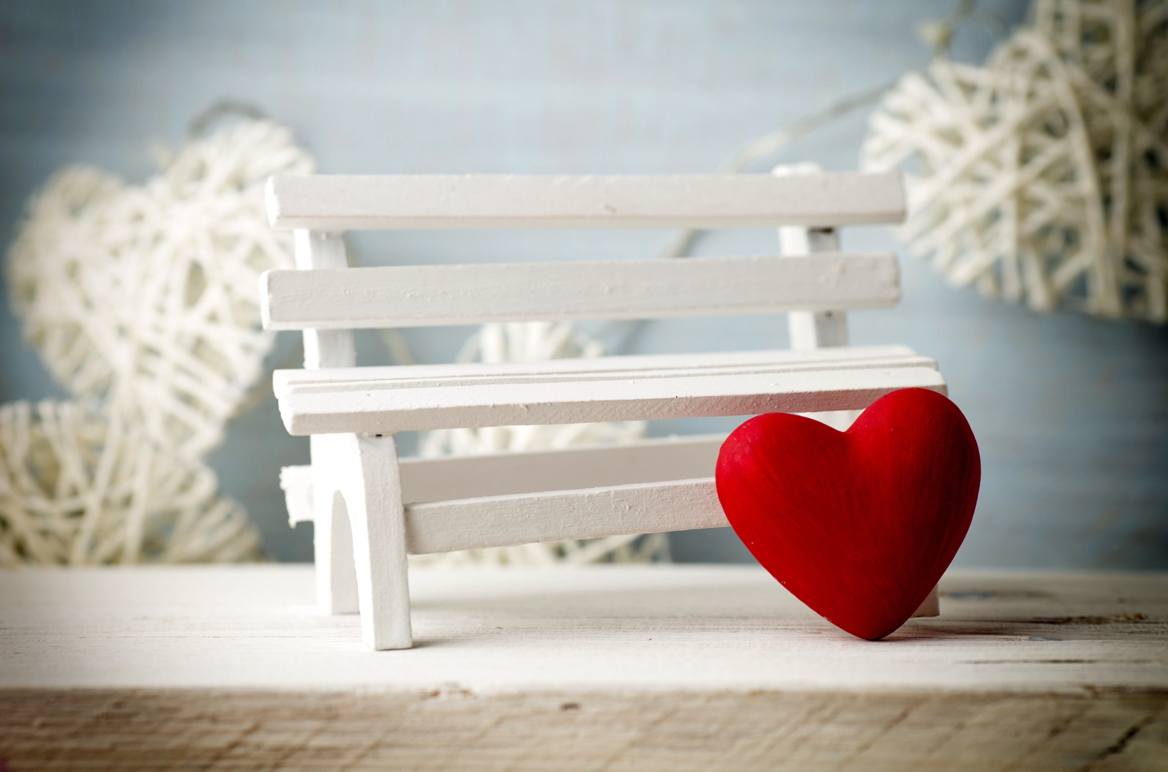 A miniature white bench with a red heart leaning against it, set against a backdrop of blurred white wicker hearts, evoking a romantic Valentine's Day scene