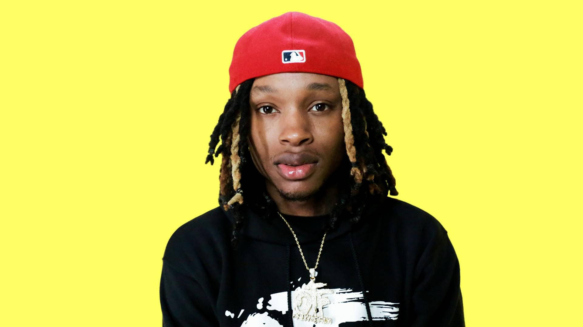 A portrait of a young man with dreadlocks, wearing a red baseball cap and a black hoodie, against a bright yellow background