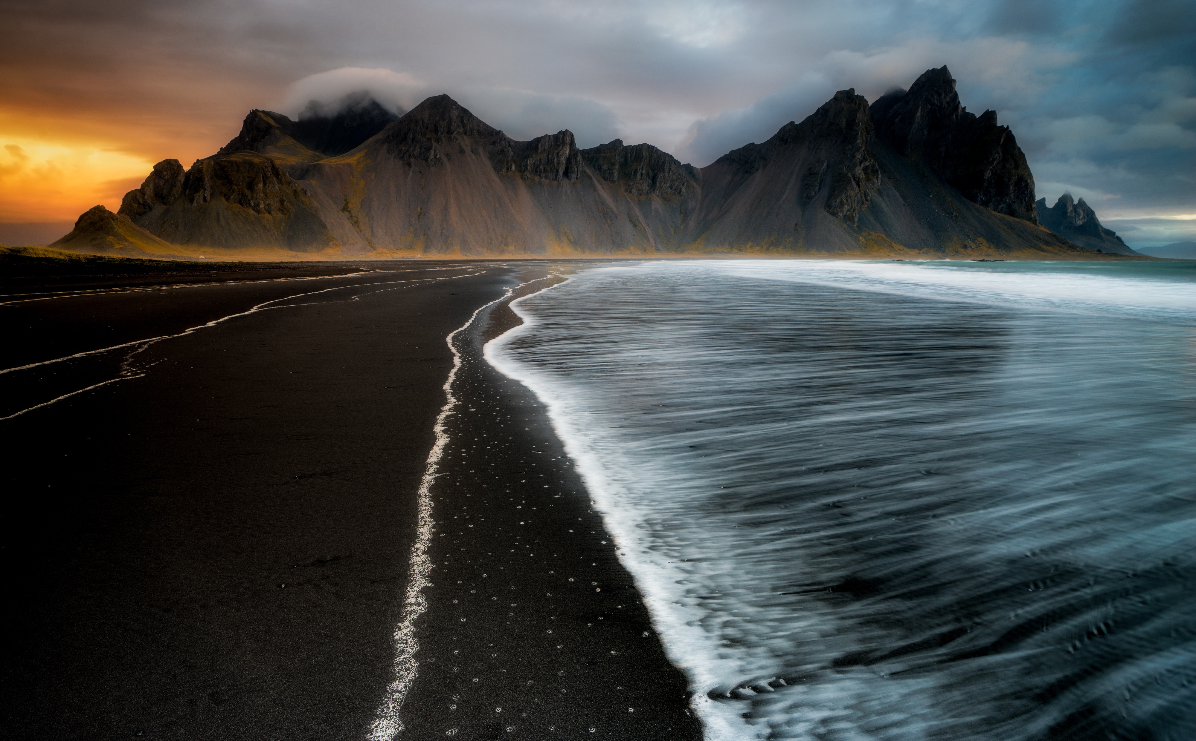 A breathtaking 4K wallpaper showcasing the dramatic beauty of a black sand beach at sunset. The smooth, dark sand contrasts with the foamy white waves, while majestic mountains rise in the background under a vibrant sky