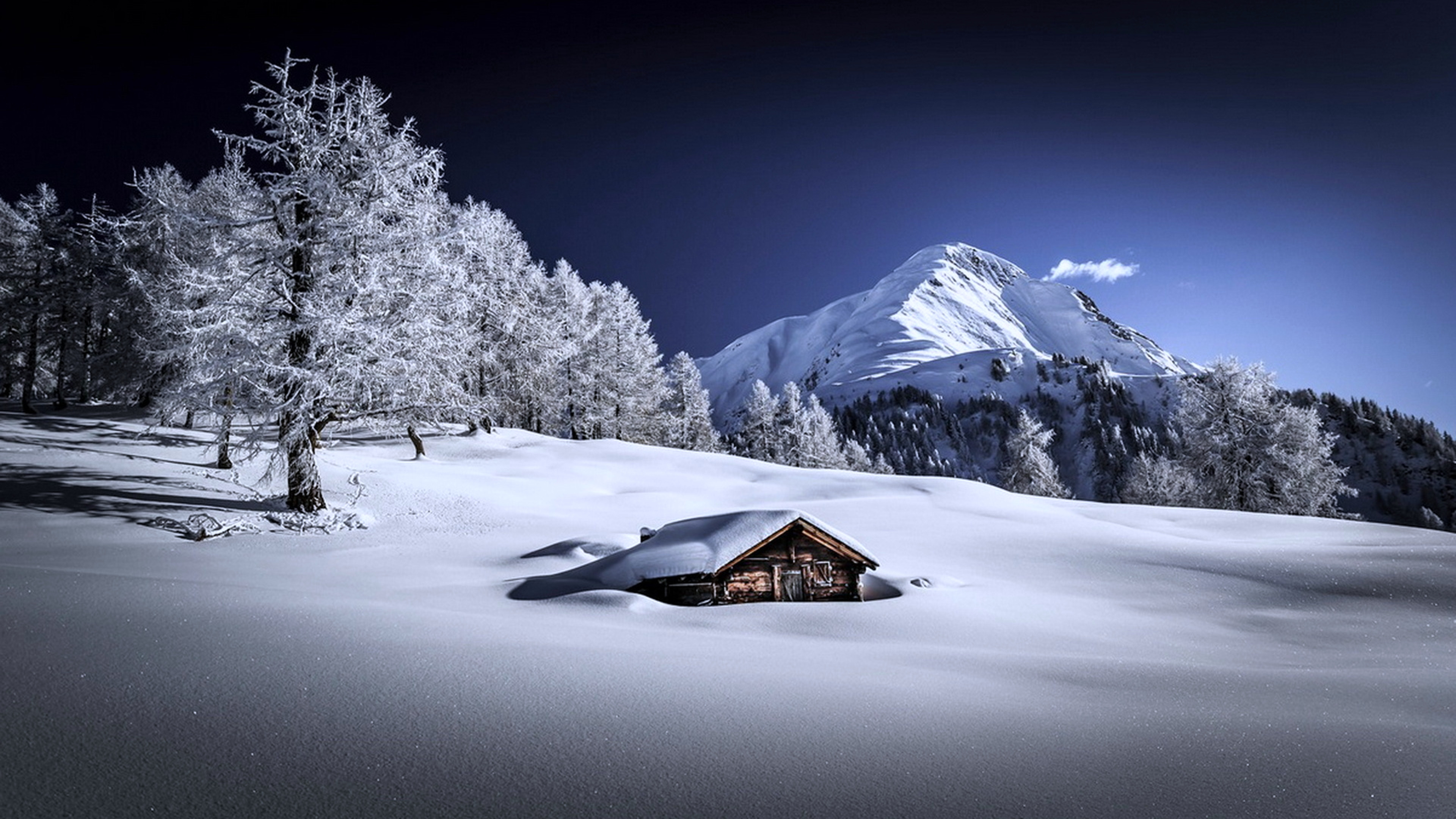A breathtaking 4k winter wallpaper featuring a snow-covered cabin nestled amidst frost-covered trees, with a majestic snow-capped mountain in the background under a clear blue sky