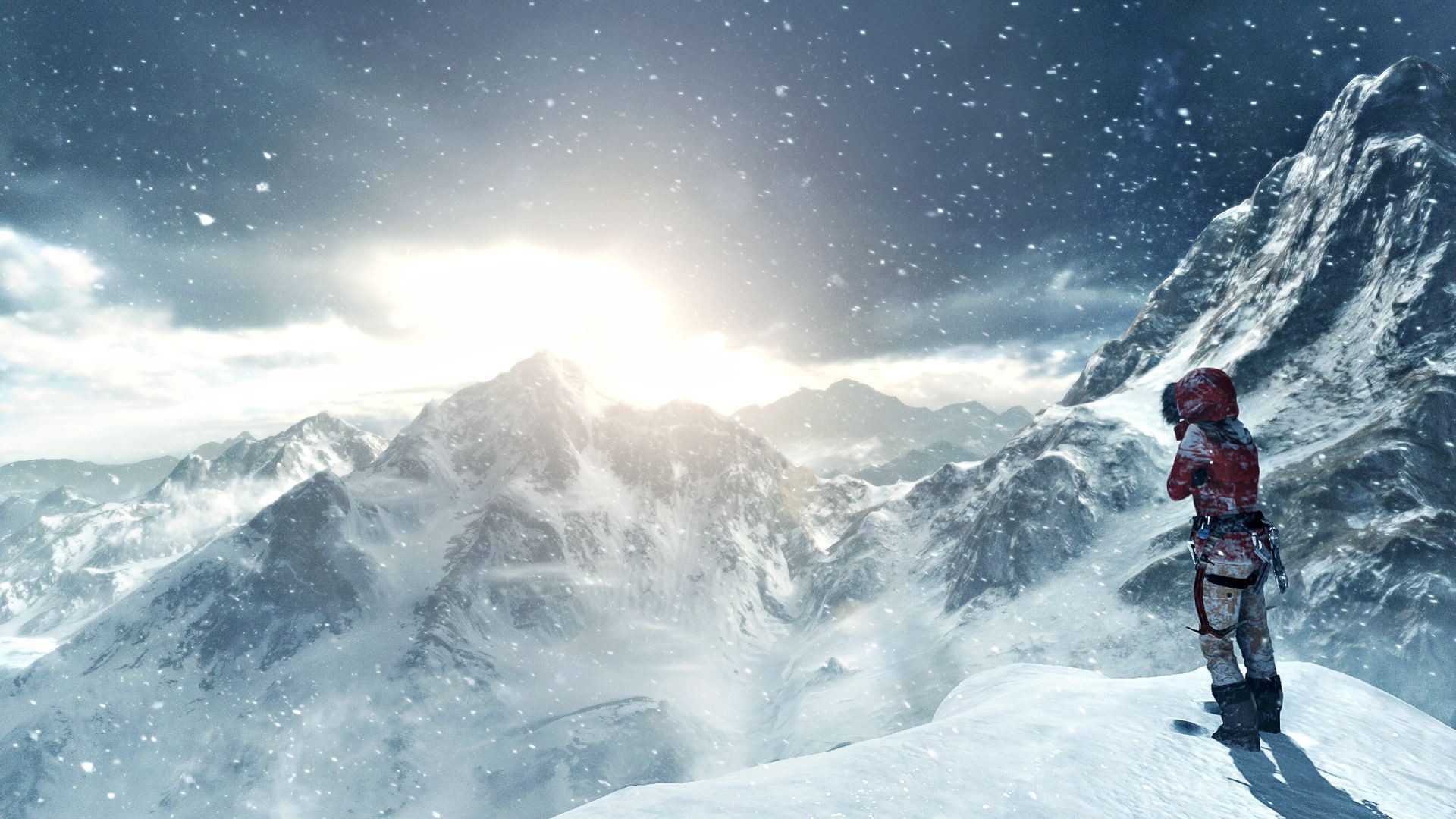 A climber in red winter gear stands on a snowy mountain peak, gazing at the vast, snow-covered mountain range under a bright, partially obscured sun. Snowflakes are falling around them