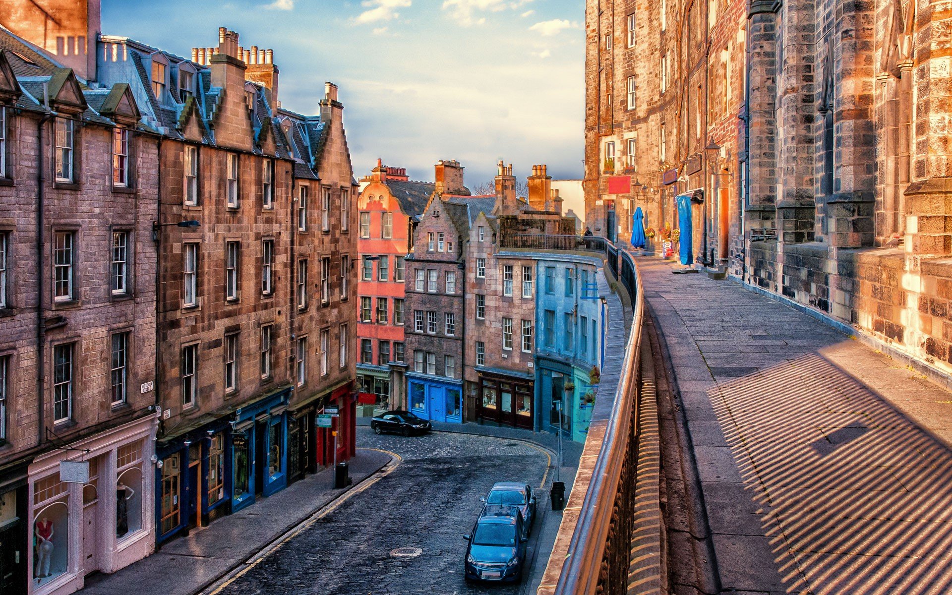 A picturesque cobblestone street in Edinburgh, Scotland, lined with colorful historic buildings. The warm glow of the late afternoon sun casts long shadows on the walkway, creating a cozy and inviting atmosphere