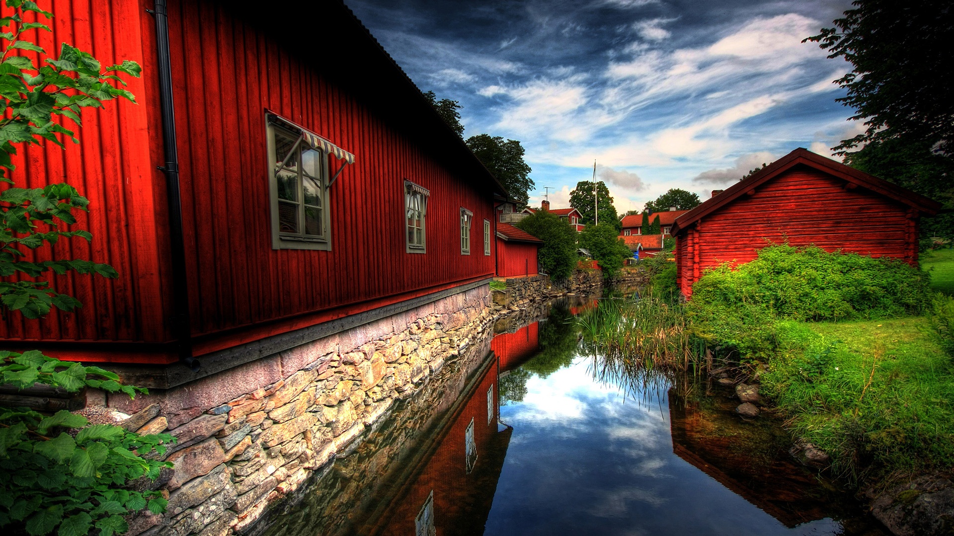 Scenic view of red wooden houses by a canal, reflecting in the water under a cloudy sky
