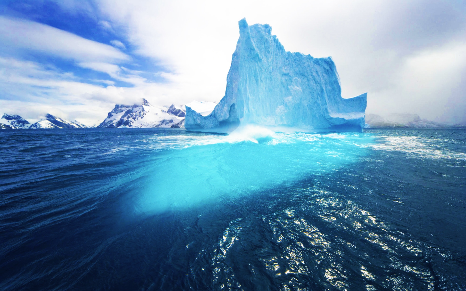 A majestic iceberg floats in the ocean, its deep blue base submerged beneath the waves. Snowy mountains create a dramatic backdrop under a cloudy sky