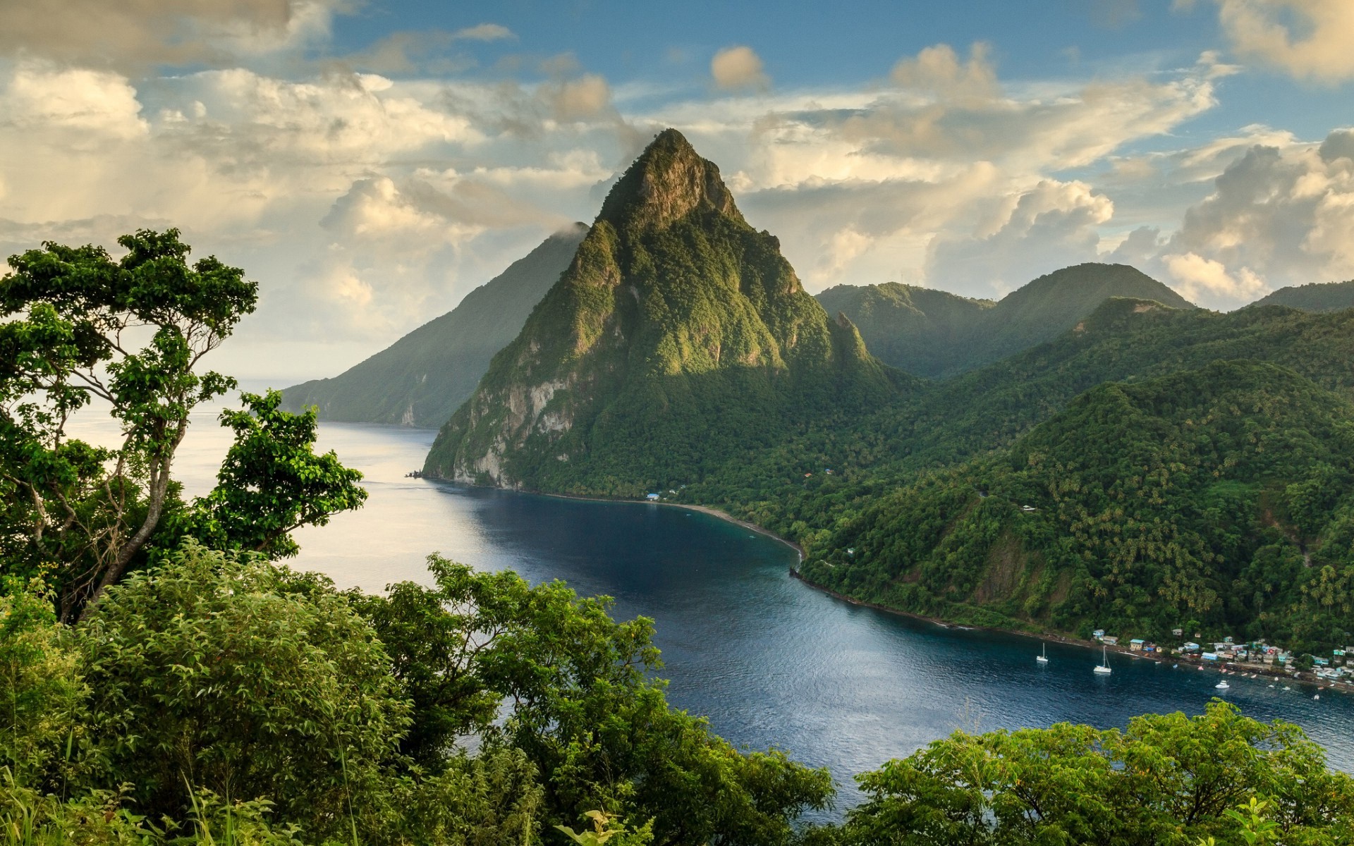 Lush green mountains meet the deep blue sea in a breathtaking tropical landscape, with a prominent peak reaching towards a cloudy sky