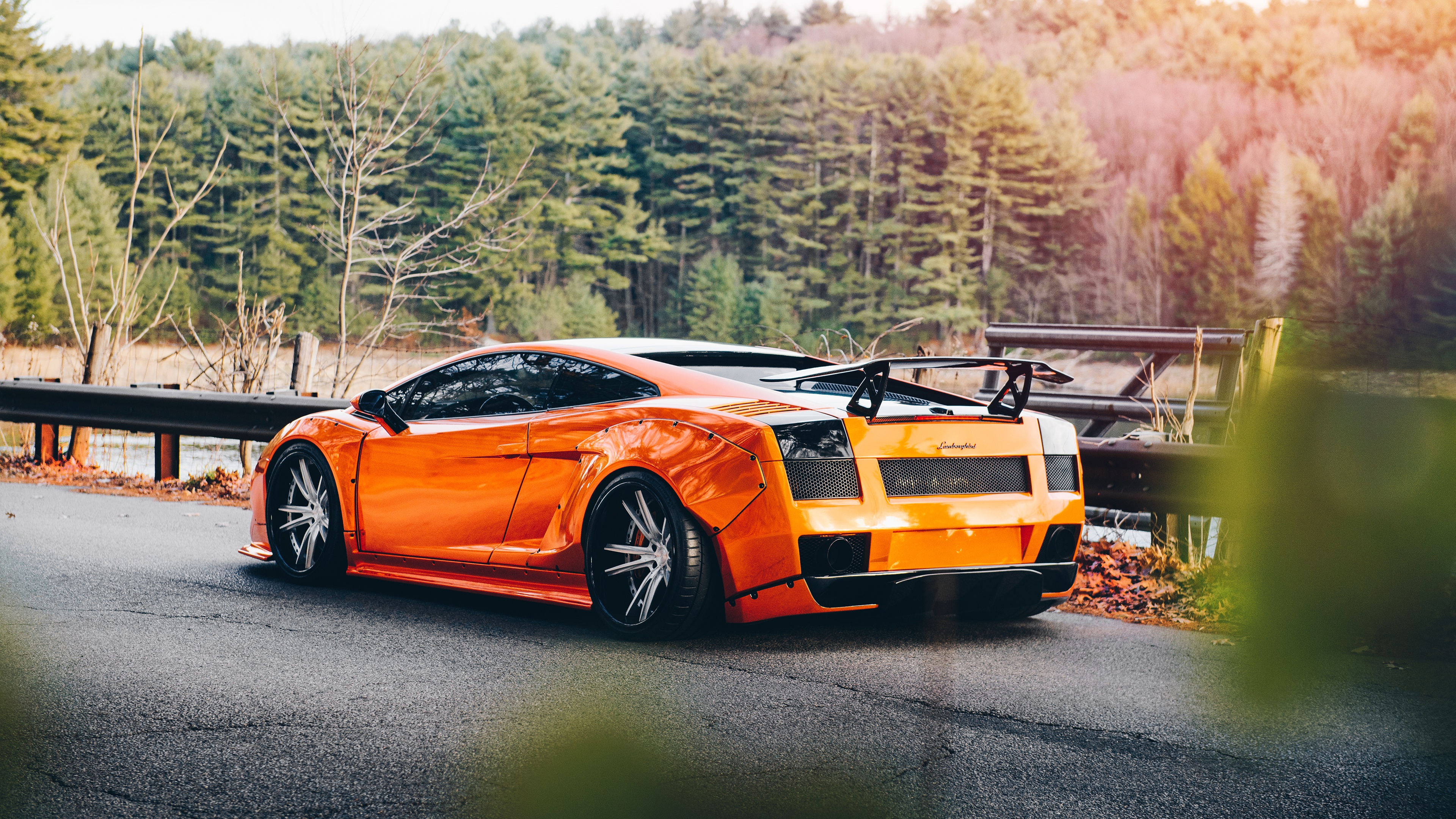 A vibrant orange Lamborghini Gallardo, customized with aftermarket body kit and wheels, parked on a scenic roadside overlooking a tranquil lake