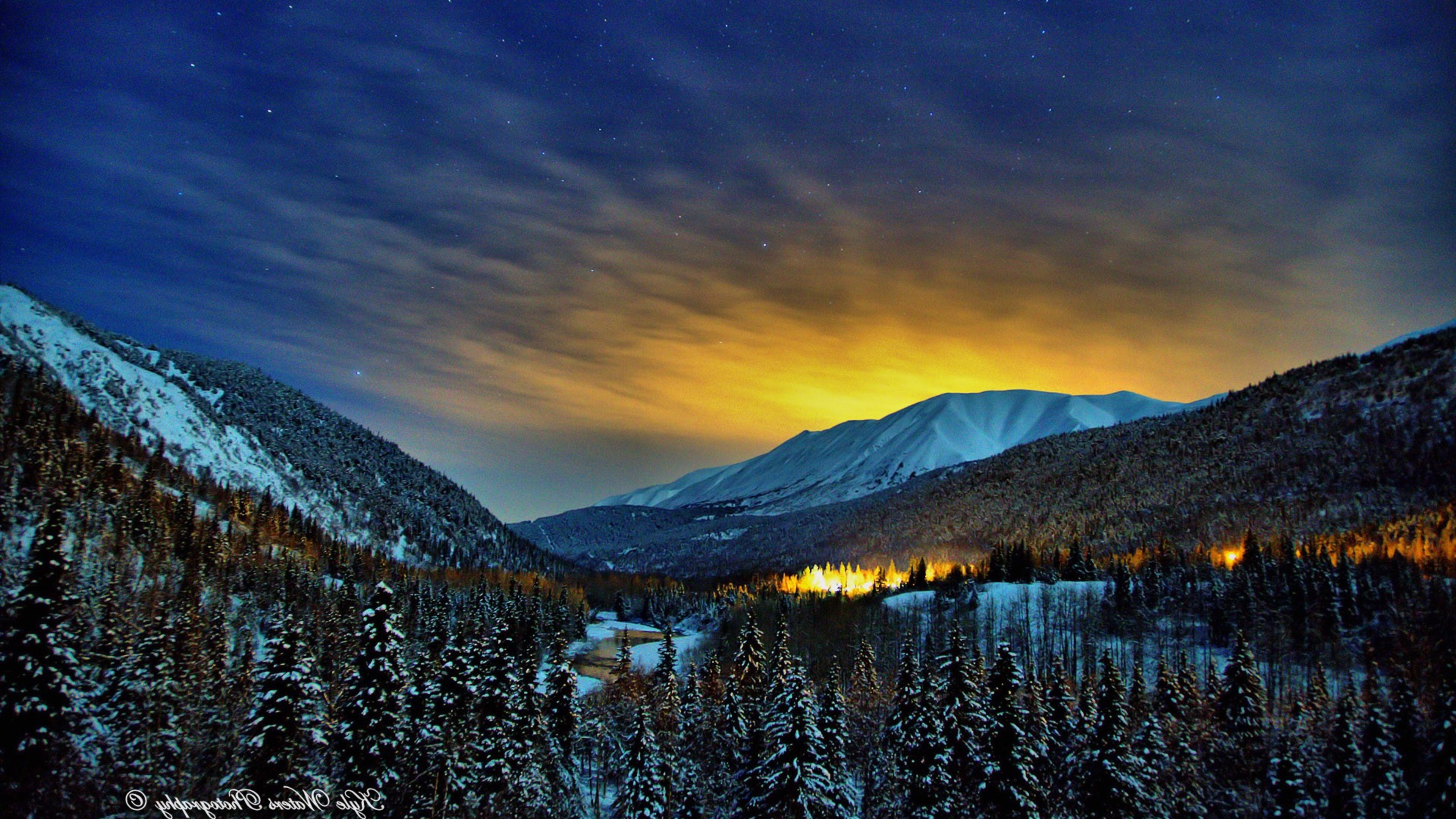 A breathtaking winter landscape at twilight, with snow-covered mountains, a serene river, and a starry sky