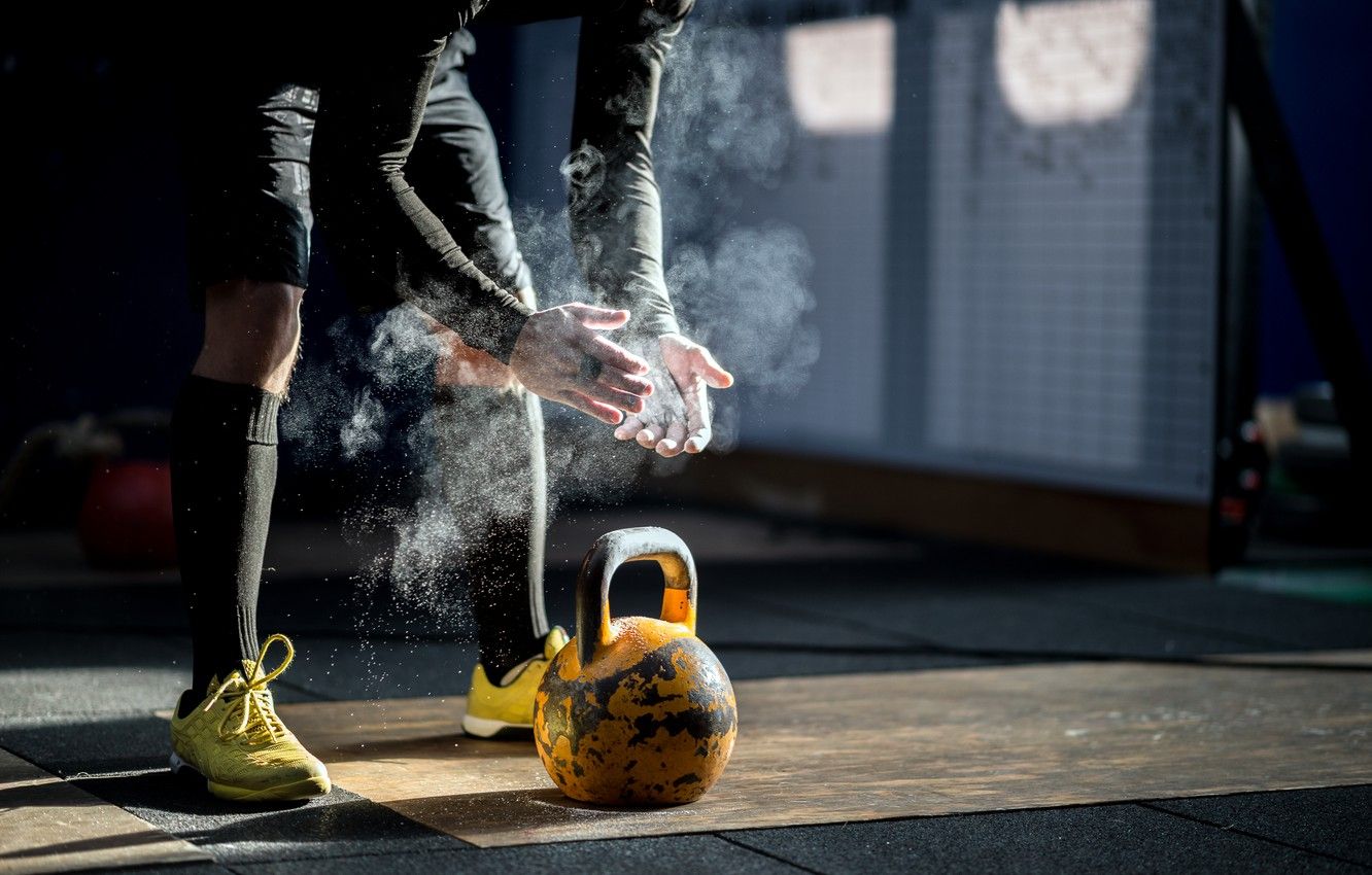 A CrossFit athlete chalks their hands, preparing to lift a kettlebell in a gym. The focus is on the athlete's hands and the kettlebell, highlighting the preparation and intensity of the workout