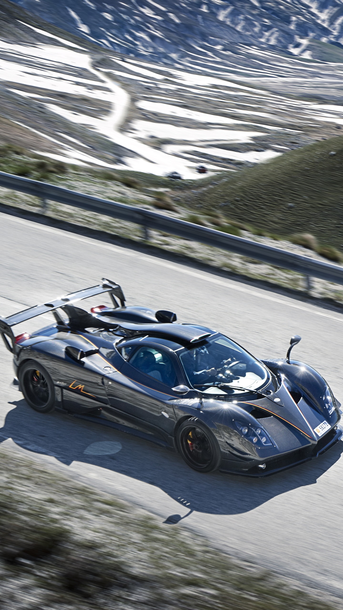 A sleek, dark gray Pagani Huayra supercar drives along a winding mountain road. Patches of snow are visible in the background. The image is perfect for Pagani enthusiasts