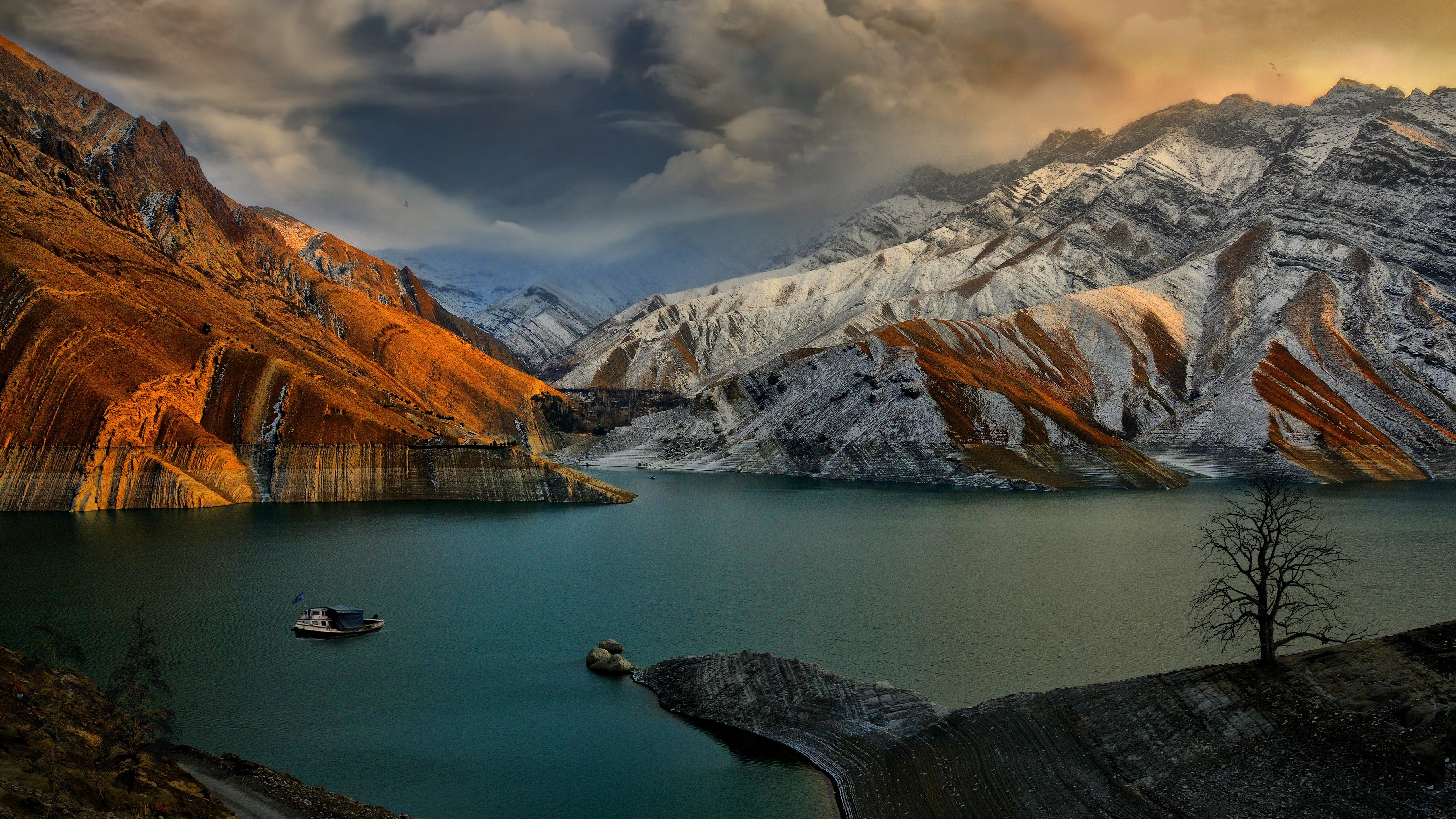 A breathtaking 5K HD wallpaper showcasing a serene lake nestled amidst snow-capped mountains. A lone boat drifts on the tranquil water, adding a touch of serenity to the dramatic landscape