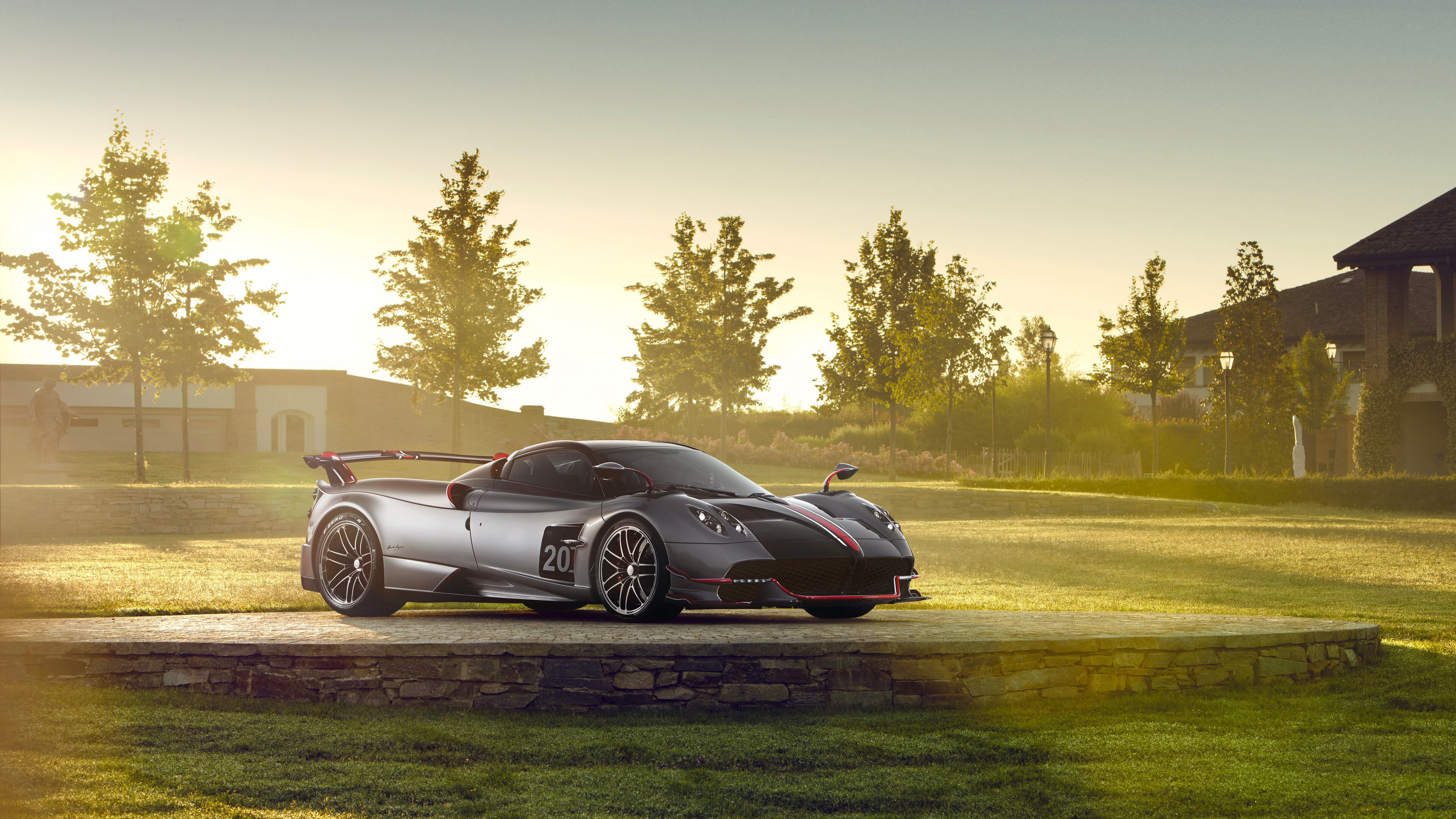 A sleek silver Pagani Huayra Roadster BC sits on a paved circle in a grassy field, bathed in the golden light of the setting sun
