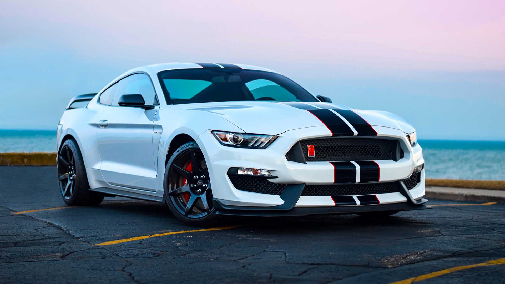 White Ford Mustang Shelby GT350 parked by the sea, featuring black racing stripes and black alloy wheels