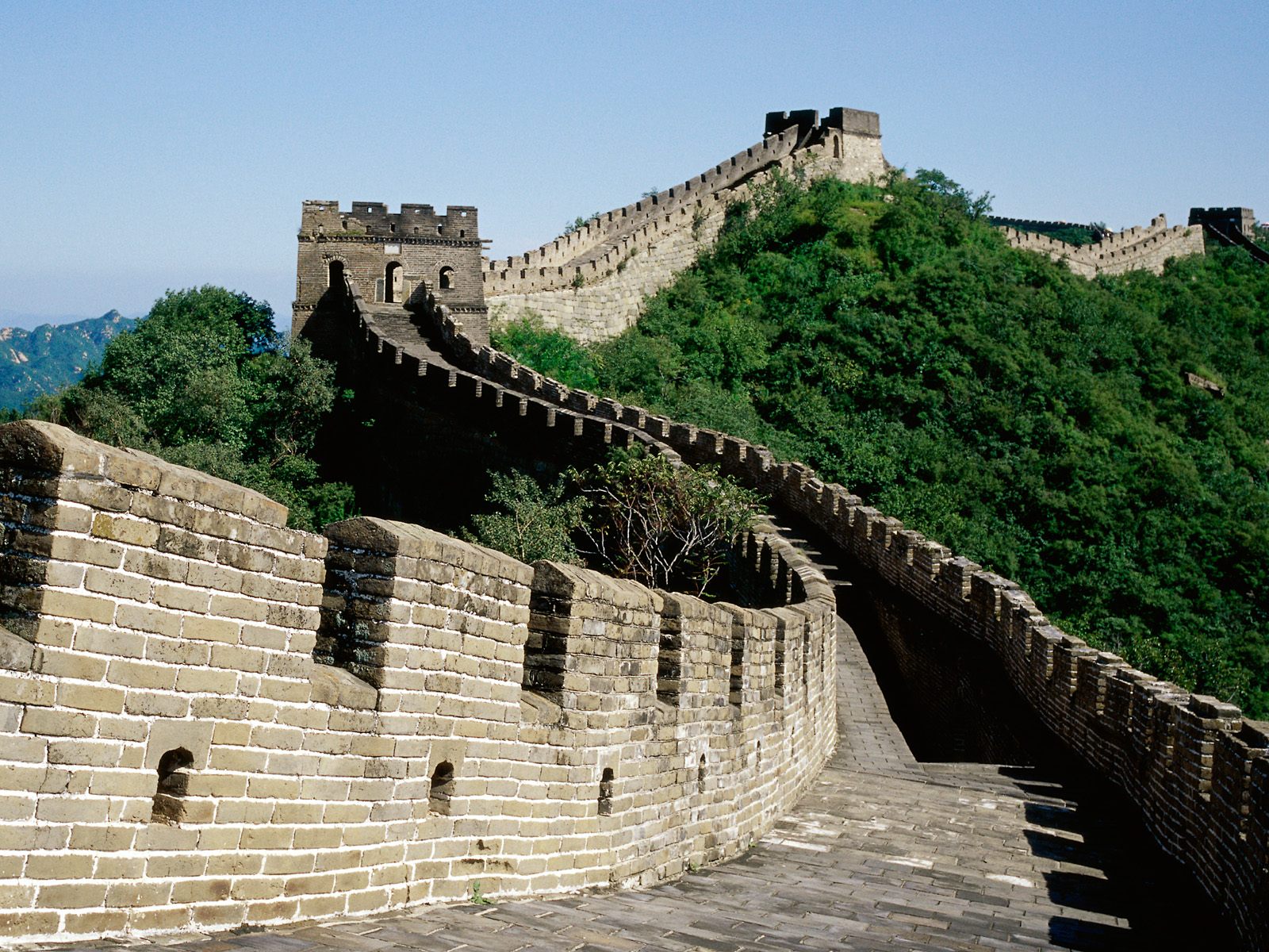 The Great Wall of China winds through lush green hills under a clear blue sky, a testament to ancient Chinese engineering and a popular tourist destination