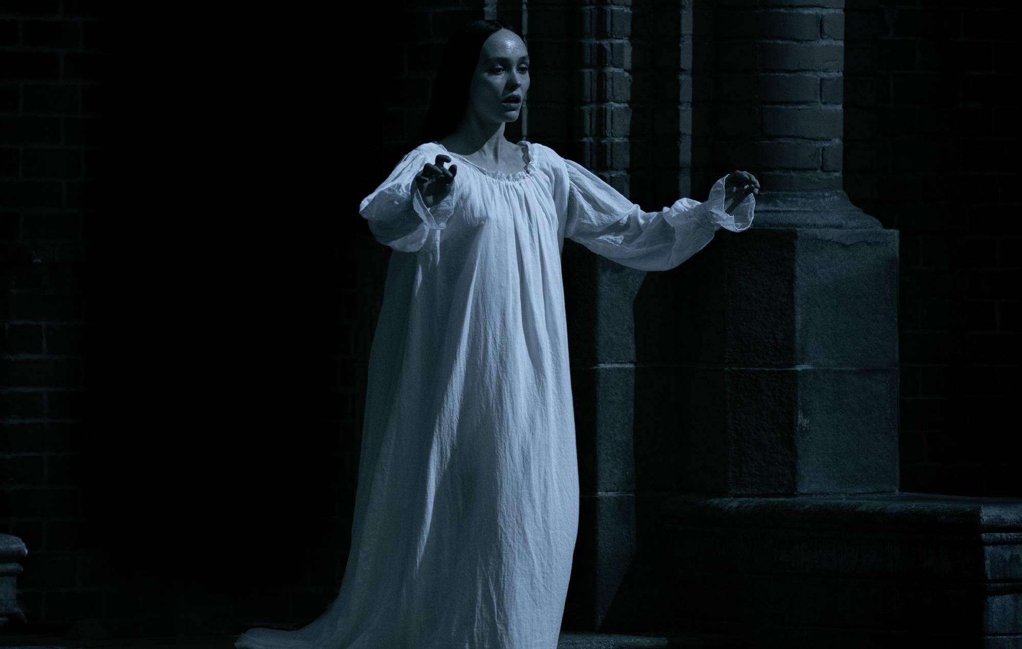 A woman in a white nightgown, reminiscent of classic vampire films, stands in a dark, gothic setting, possibly a church or castle, with a look of fear or apprehension. Her pose suggests she might be sleepwalking or under some supernatural influence
