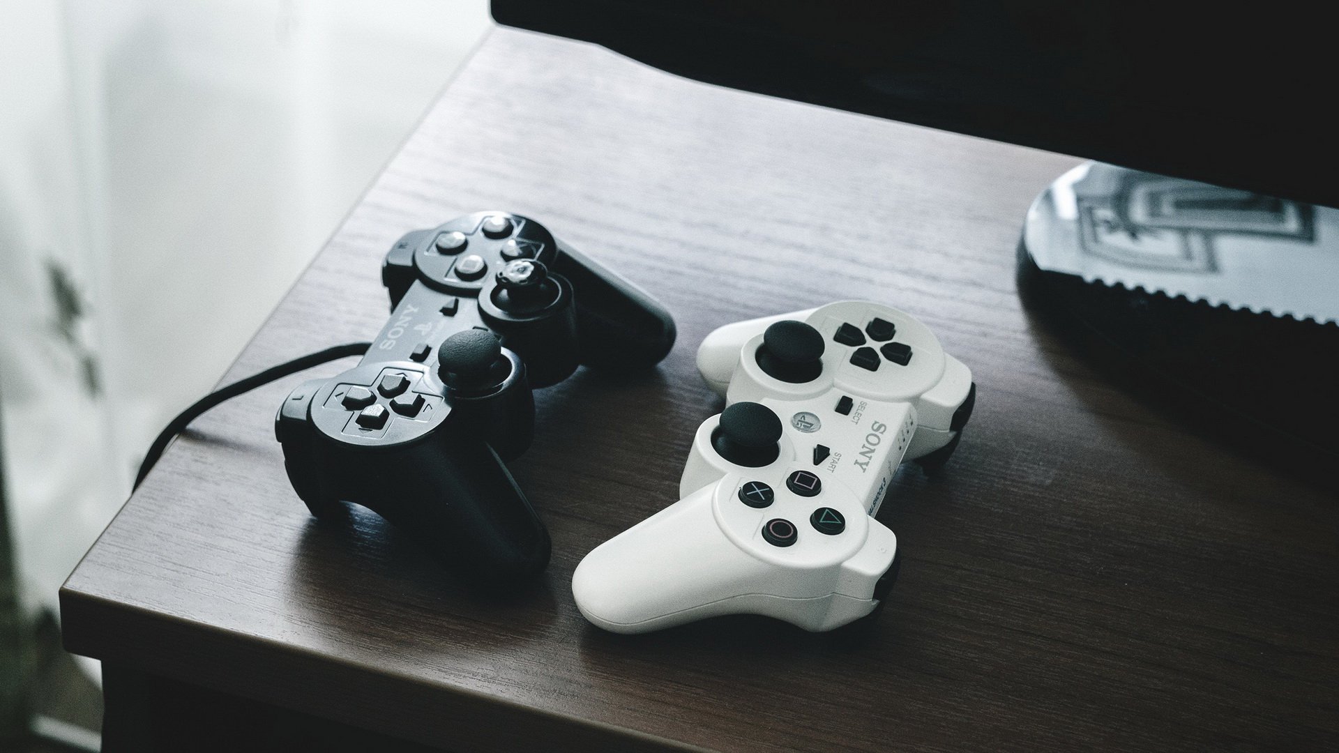 Two PlayStation controllers, one black and one white, rest on a wooden table in front of a blurred television screen